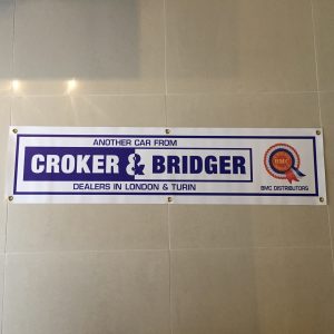 BMC CROKER AND BRIDGER BANNER. A white banner with brass eyelets. Croker & Bridger in blue and white lettering next to a BMC rosette. Additional text in blue; Another Car From Dealers In London & Turin; BMC Distributors. All bordered in blue.