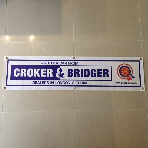 A white banner with brass eyelets. Croker & Bridger in blue and white lettering next to a BMC rosette. Additional text in blue; Another Car From Dealers In London & Turin; BMC Distributors. All bordered in blue.