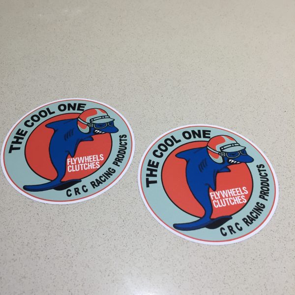 CRC RACING PRODUCTS STICKERS. The Cool One CRC Racing Products in black lettering surrounds a blue circle. In the centre on a red circle is a blue shark wearing a red and blue scooter helmet and goggles next to Flywheels Clutches in white lettering.