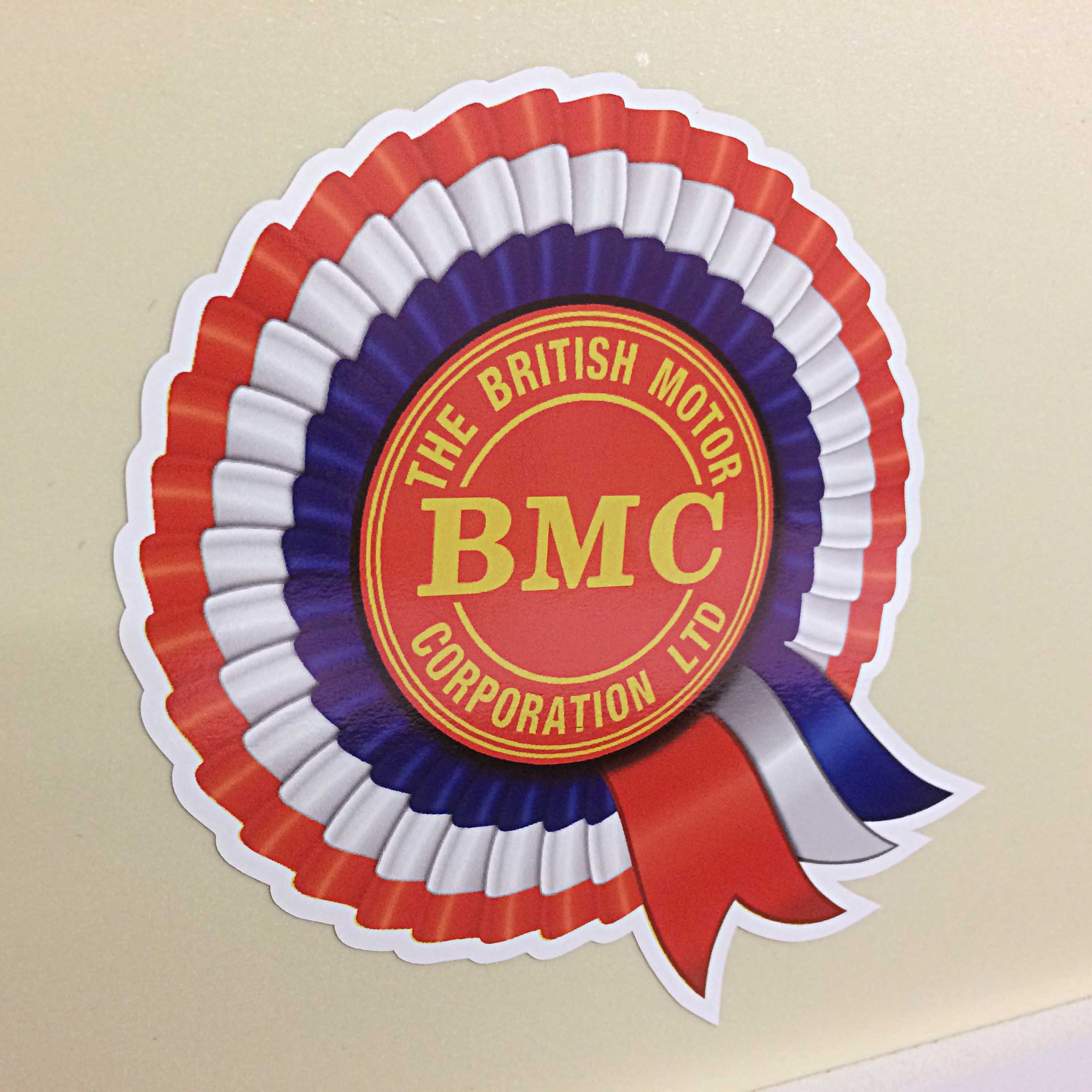 BMC ROSETTE STICKER. BMC The British Motor Corporation Ltd in yellow uppercase lettering surrounds a red disc in the centre of a red, white and blue rosette.