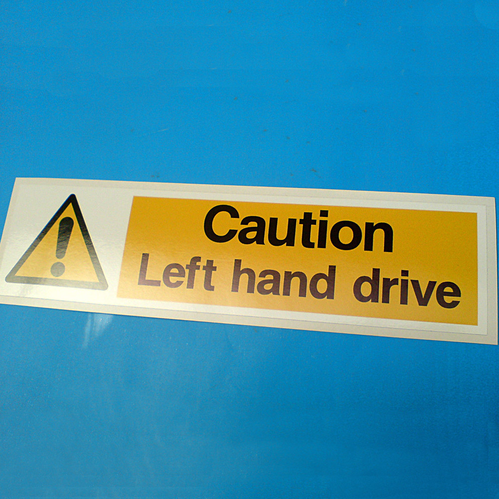 A white rectangular sticker. On the left is a yellow triangle with a black border and a black exclamation mark in the centre. Next to this Caution Left hand drive in black lettering within a yellow rectangle.