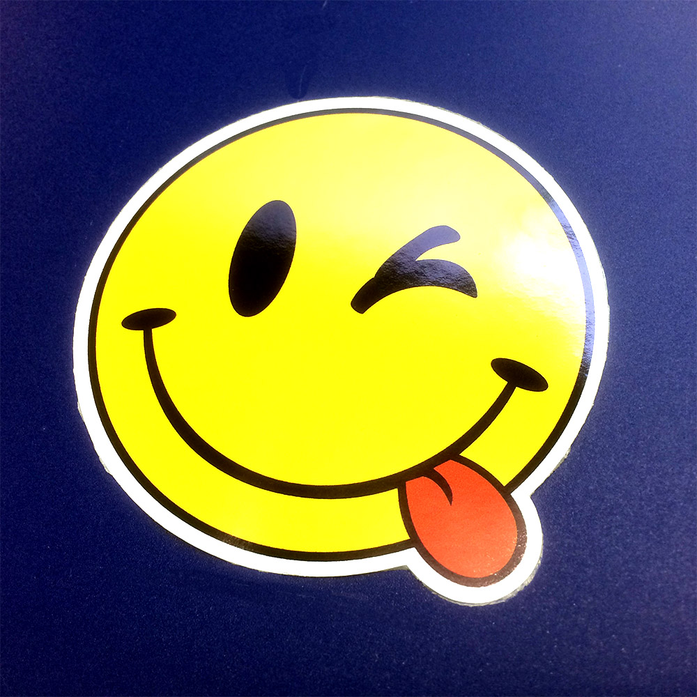 A round yellow smiling face. The eyes and mouth are black. It is pulling a red tongue and winking an eye.