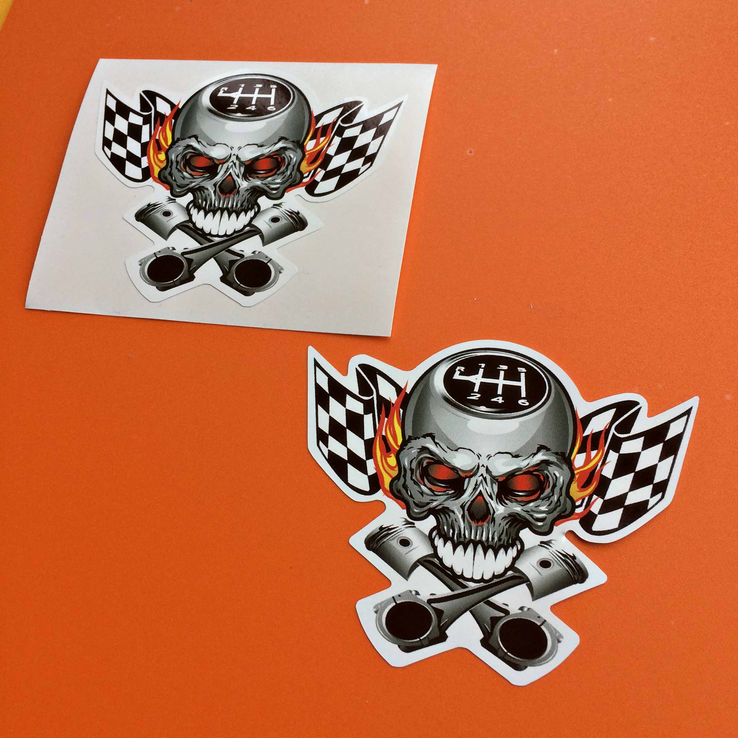 CHEQUERED SKULL AND PISTONS STICKERS. Two crossed pistons below a silver skull with red eyes and white teeth. On the scalp of the skull are the symbols R 1 2 3 4 5 6 as on a car gearstick. A chequered flag and flames are in the background.