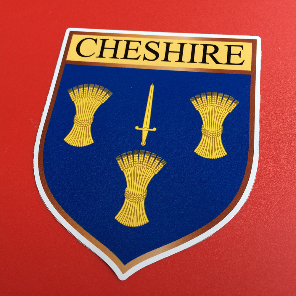 CHESHIRE COUNTY SHIELD STICKER. Cheshire in black uppercase lettering on a yellow banner across the top of a shield. The shield displays three golden sheaves of wheat and a golden blade on a blue background.