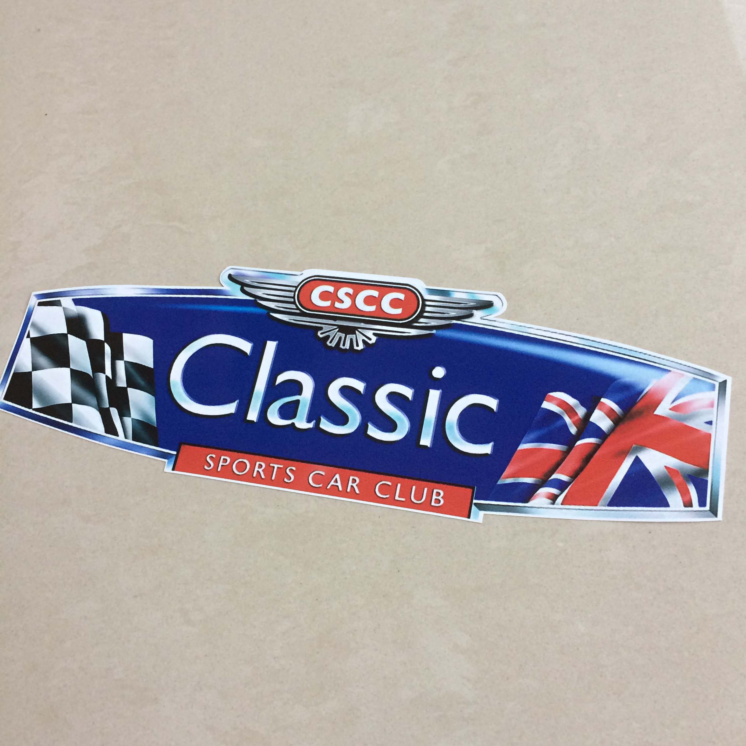 CLASSIC SPORTS CAR CLUB STICKER. A blue sticker with a silver edge imitating the grill of a vintage car with a chequered flag and a Union Jack on either side. Classic in white lettering in the centre. Sports Car Club below on a red badge. Above silver wings and CSCC on a red badge.