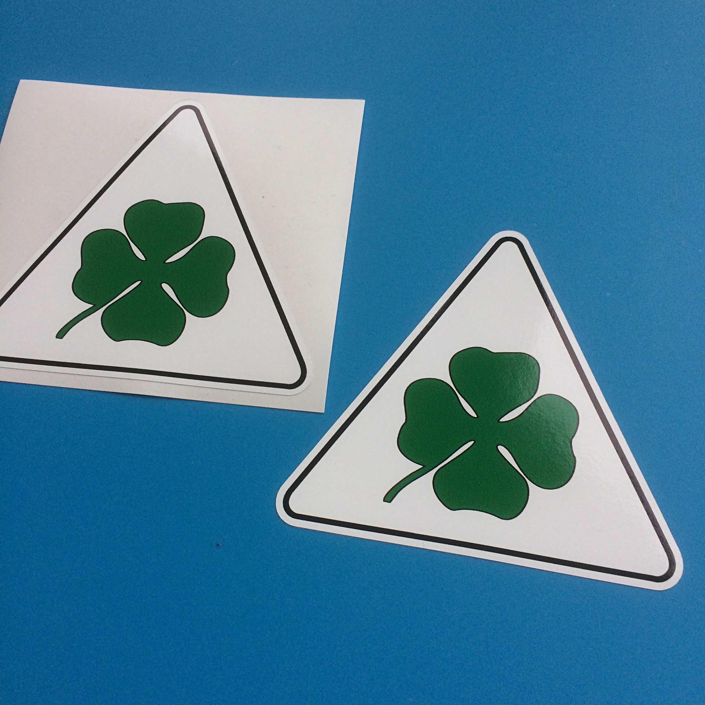 A white triangular sticker with a black border. In the centre is a green four leaf clover.