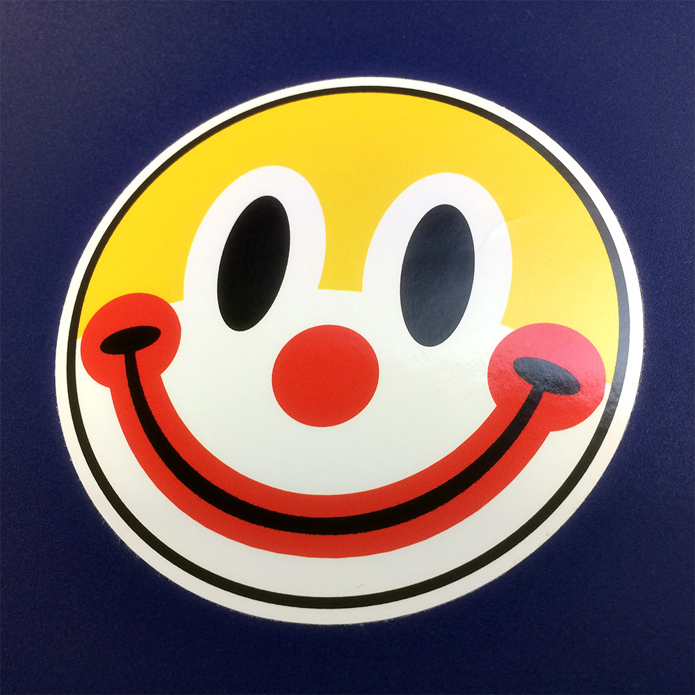 CLOWN SMILEY STICKER. A round white face of a clown with yellow hair, black eyes, a red nose and black and red smiling mouth.