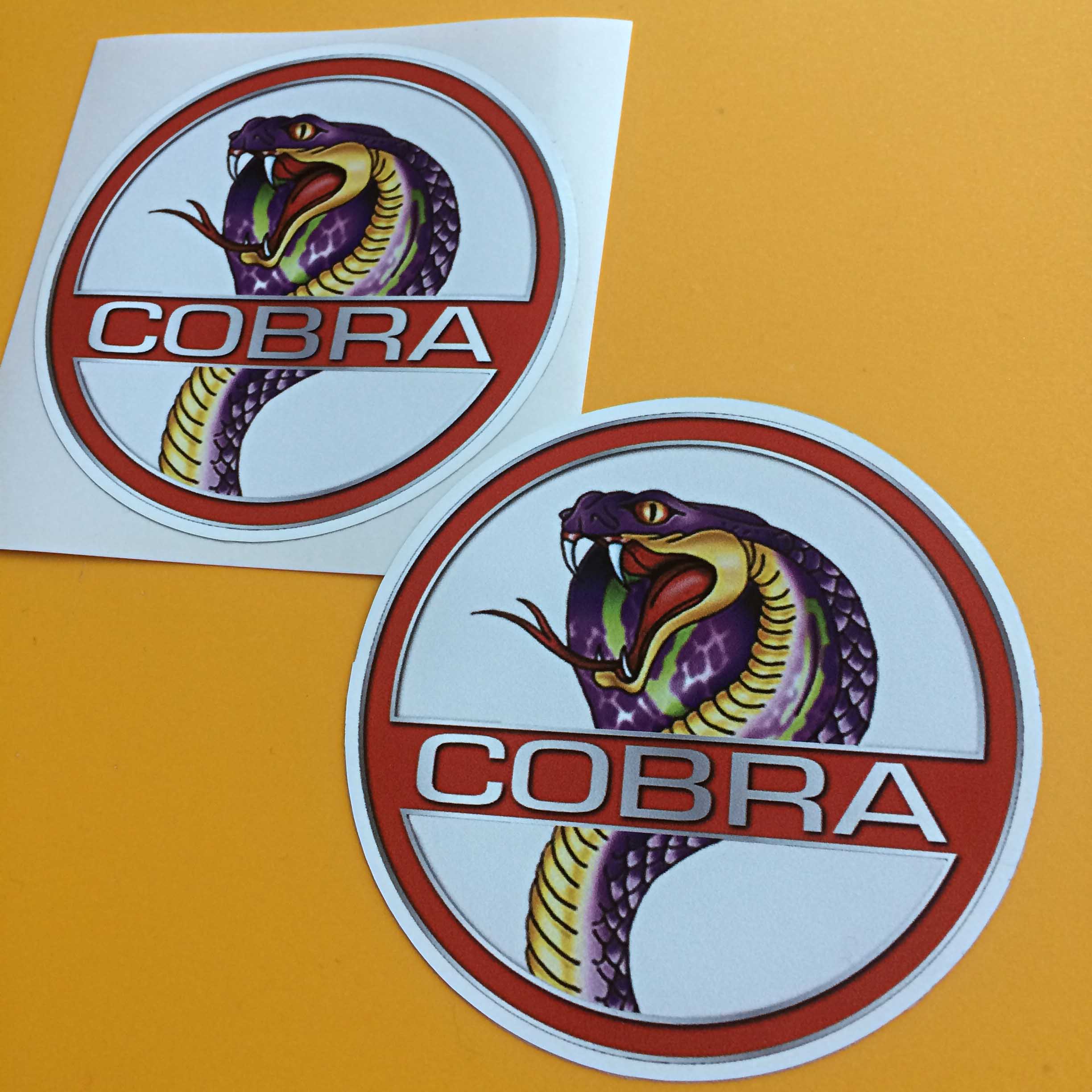 COBRA STICKERS. A white circular sticker with a red border. Across the centre on a red banner is Cobra in white uppercase lettering. In the background is the head of a Cobra in purple, yellow and green displaying a red tongue and white fangs.