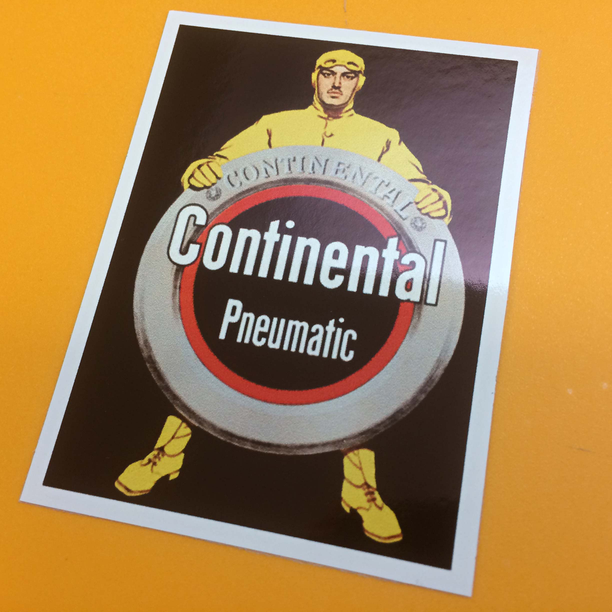 CONTINENTAL PNEUMATIC STICKER. A man wearing a yellow vintage car racing suit, goggles and ear muffs on a black background. He is standing behind and resting both hands on a Continental tyre. Continental Pneumatic in white lettering overlays the image.