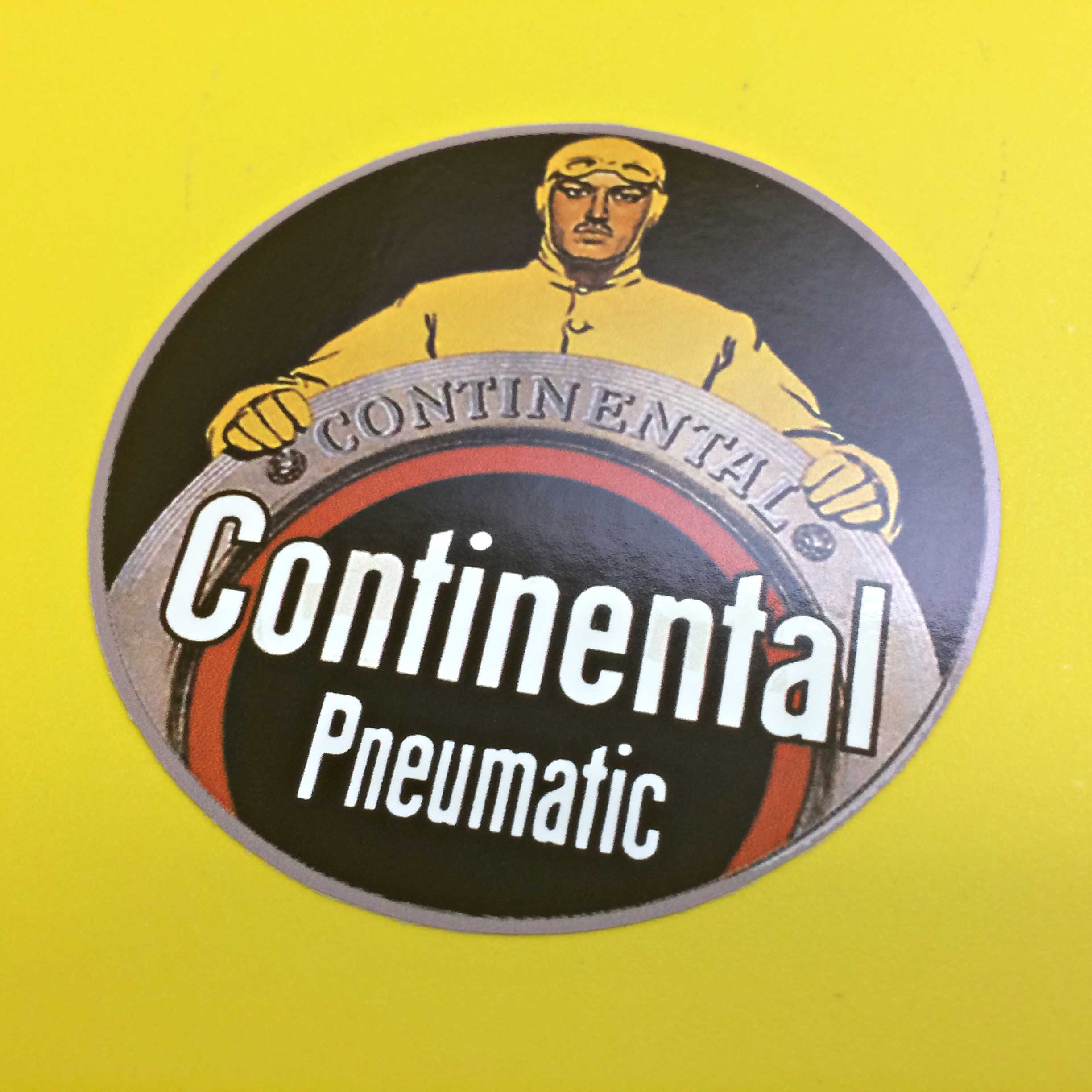 CONTINENTAL PNEUMATIC STICKER. Continental Pneumatic in white lettering on a black circular sticker. A man wearing a yellow vintage car racing suit with goggles and ear muffs is standing behind and resting both hands on a Continental tyre.