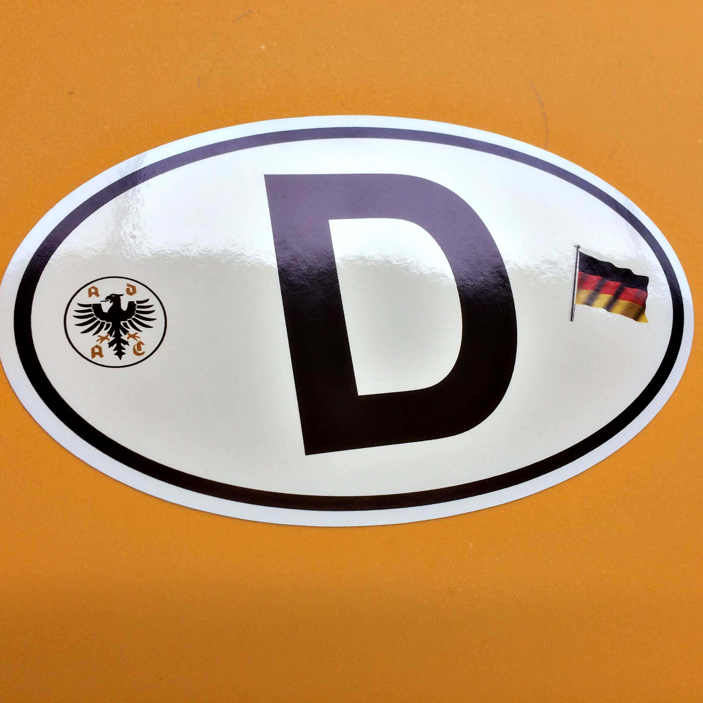 D FOR DEUTSCHLAND GERMAN STICKER. D in bold black lettering on a white oval sticker with a black border. Either side of the letter D are the German flag, three horizontal bands of black, red and gold and the ADAC, the German Automobile Club logo, a black eagle surrounded by gold letters ADAC.