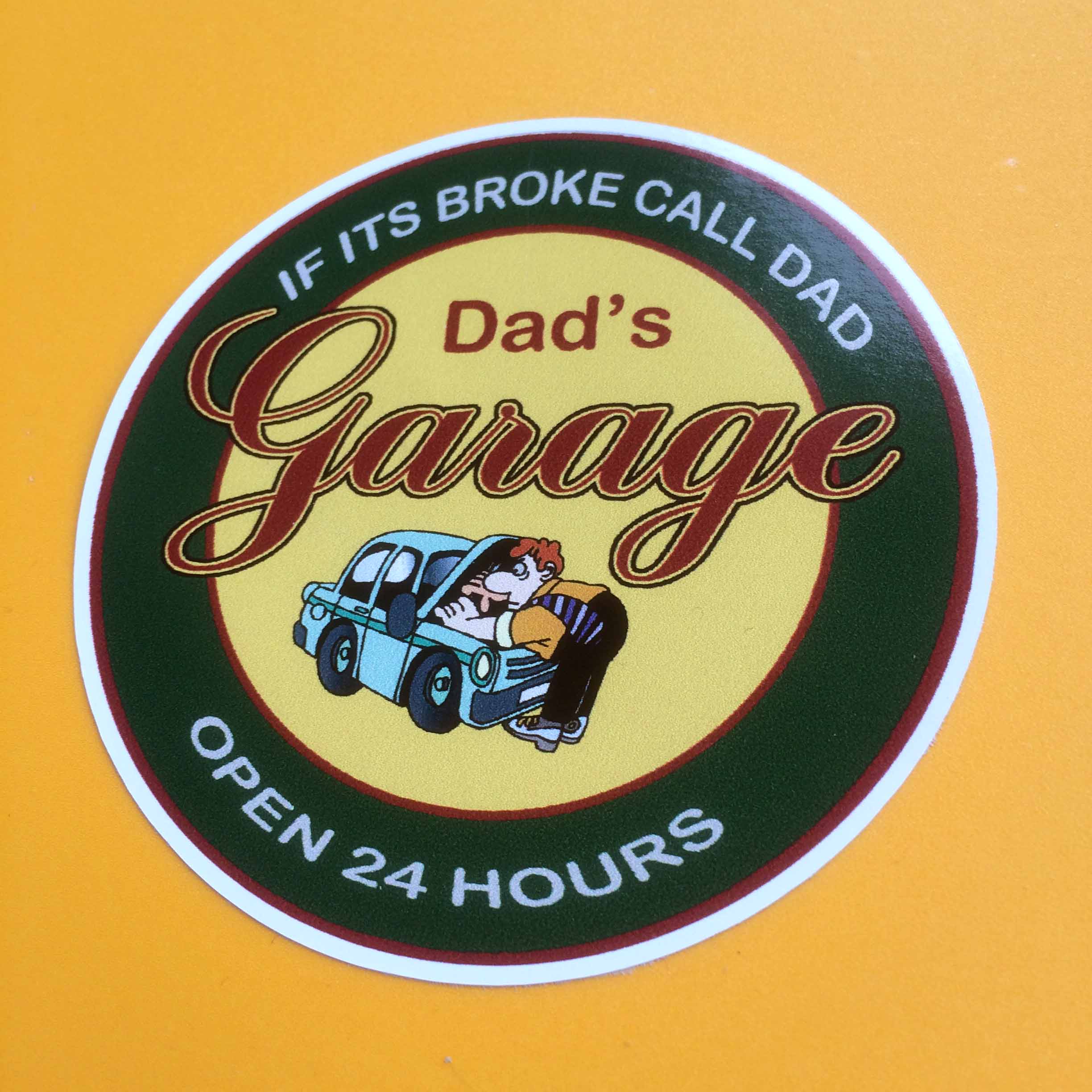 DAD'S GARAGE STICKER. Dad's Garage in burgundy lettering and a cartoon character dressed in shirt, tie and trousers working under the bonnet of a blue car on a yellow circular sticker. If it's broke call dad open 24 hours in white lettering surrounds this on a green border.
