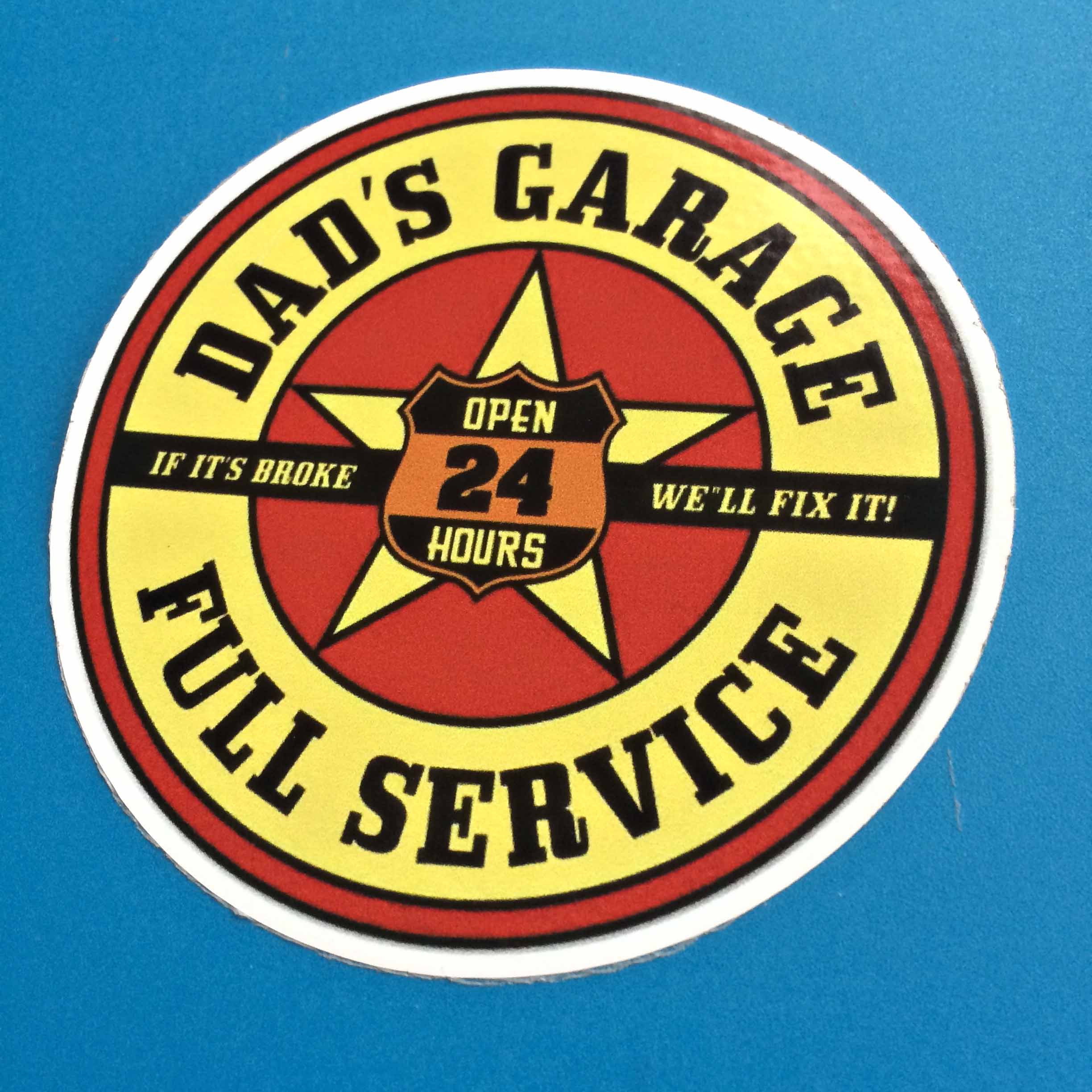 DAD'S GARAGE FULL SERVICE STICKER. Two concentric circles in yellow and red. Dad's Garage Full Service in black lettering surrounds the yellow circle. In the red circle is a yellow star overlaid with a black and orange shield, Open 24 Hours. Across the sticker is a black banner, If It's Broke We'll Fix It!