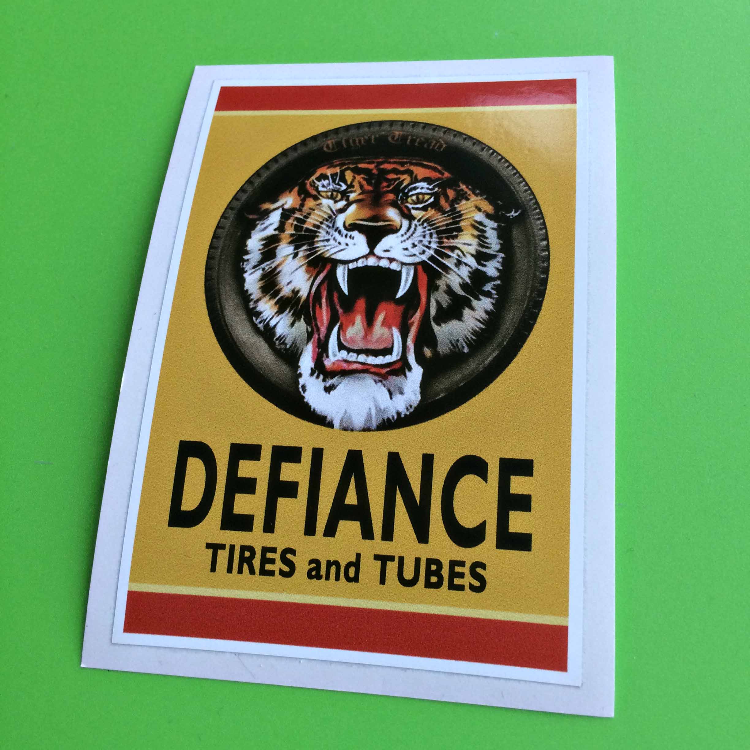 DEFIANCE TIRES AND TUBES STICKER. Defiance Tires and Tubes in black uppercase lettering on a yellow sticker with the top and bottom edges in red. A tiger's head with mouth wide open displaying a red tongue and sharp fangs sits inside a tyre.