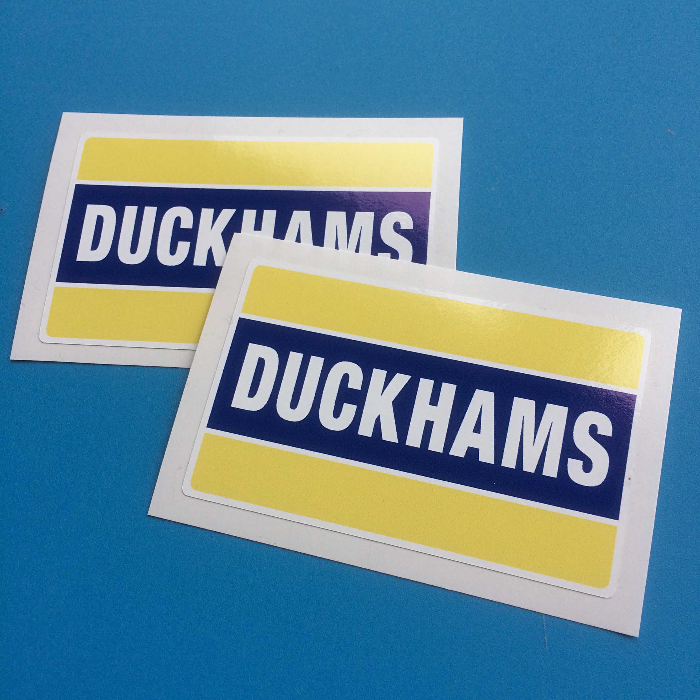 DUCKHAMS MOTOR OIL STICKERS. Duckhams in white capital letters on blue across the centre. The area above and below is yellow.