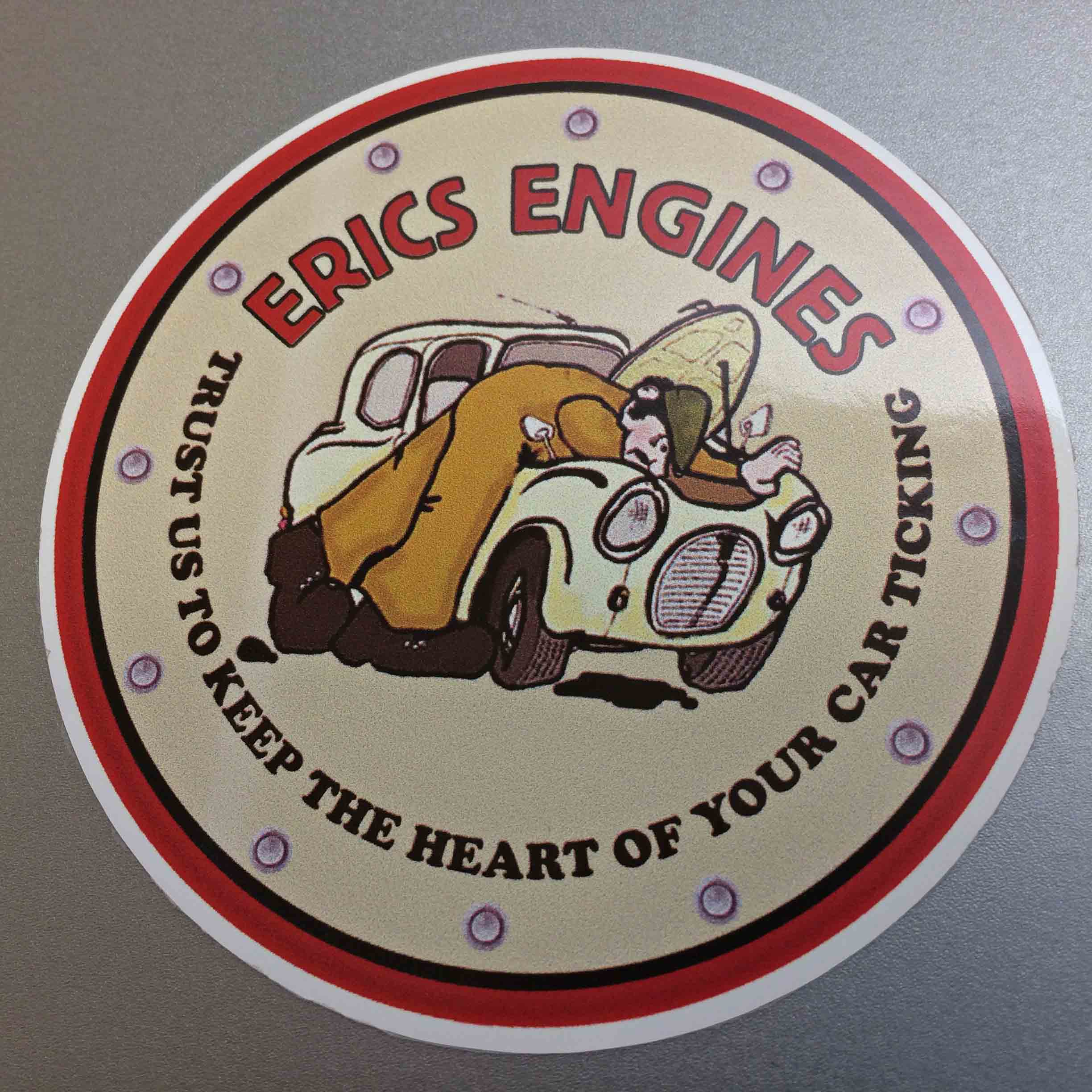 Erics Engines in red lettering. Trust Us To Keep The Heart Of Your Car Ticking in black lettering surrounds a circular sticker with a red border. In the centre is a humorous man wearing a brown boiler suit and a green flat cap working under the bonnet of a car.