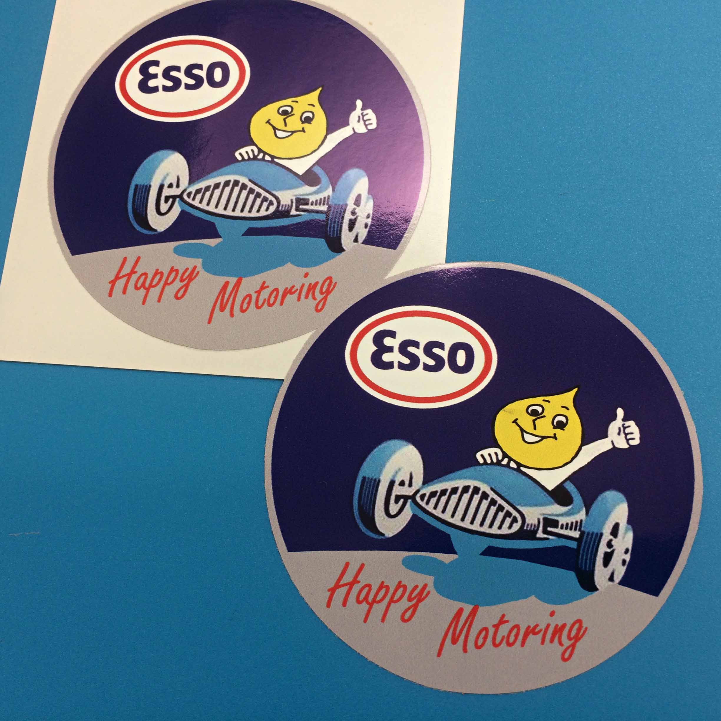 ESSO HAPPY MOTORING STICKERS. The Esso Oil Drop Man driving a blue sports car while giving the thumbs up. The Esso logo and Happy Motoring in red lettering is above and below.