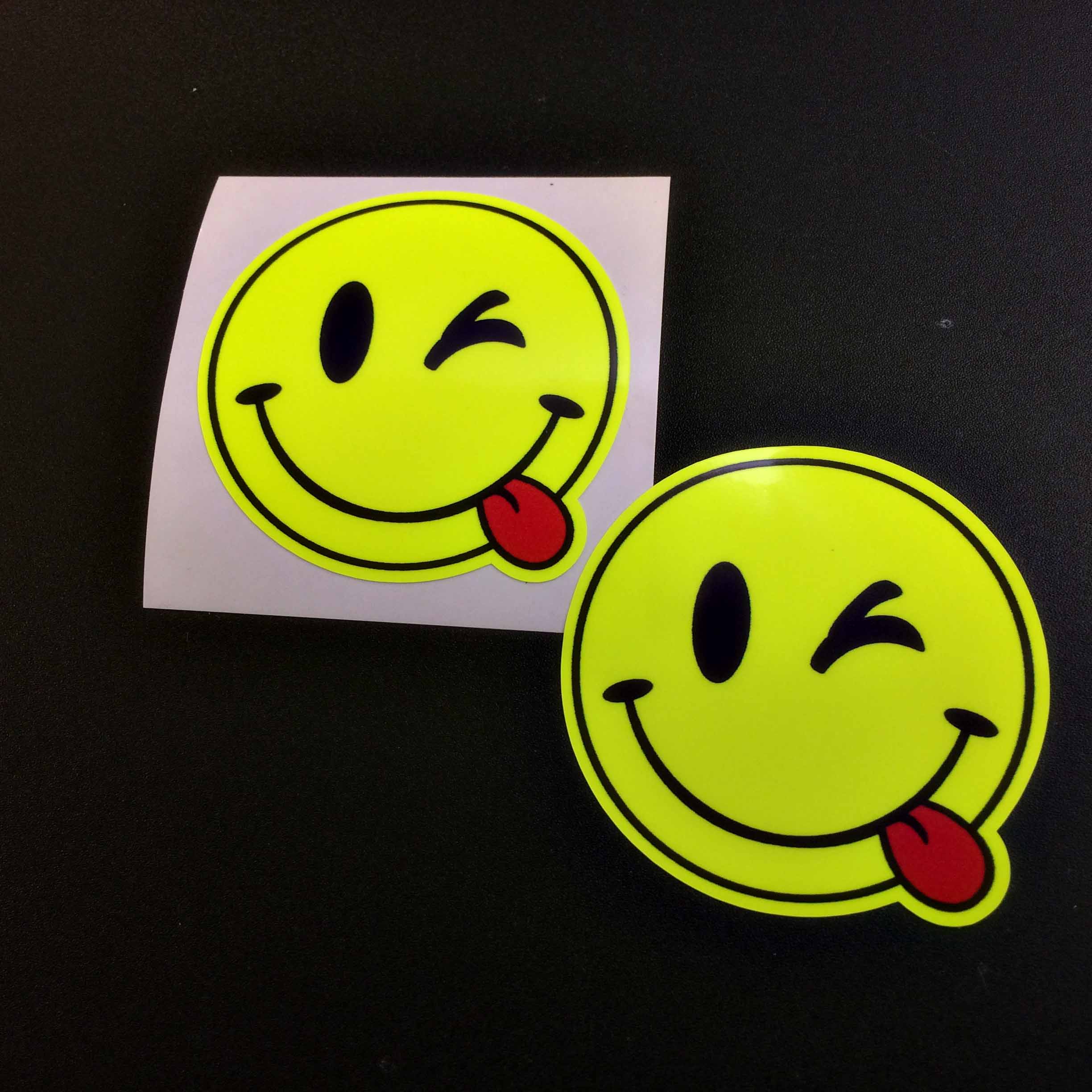 A fluorescent yellow round smiling face. A red tongue is hanging out of the corner of the mouth and one eye is winking.
