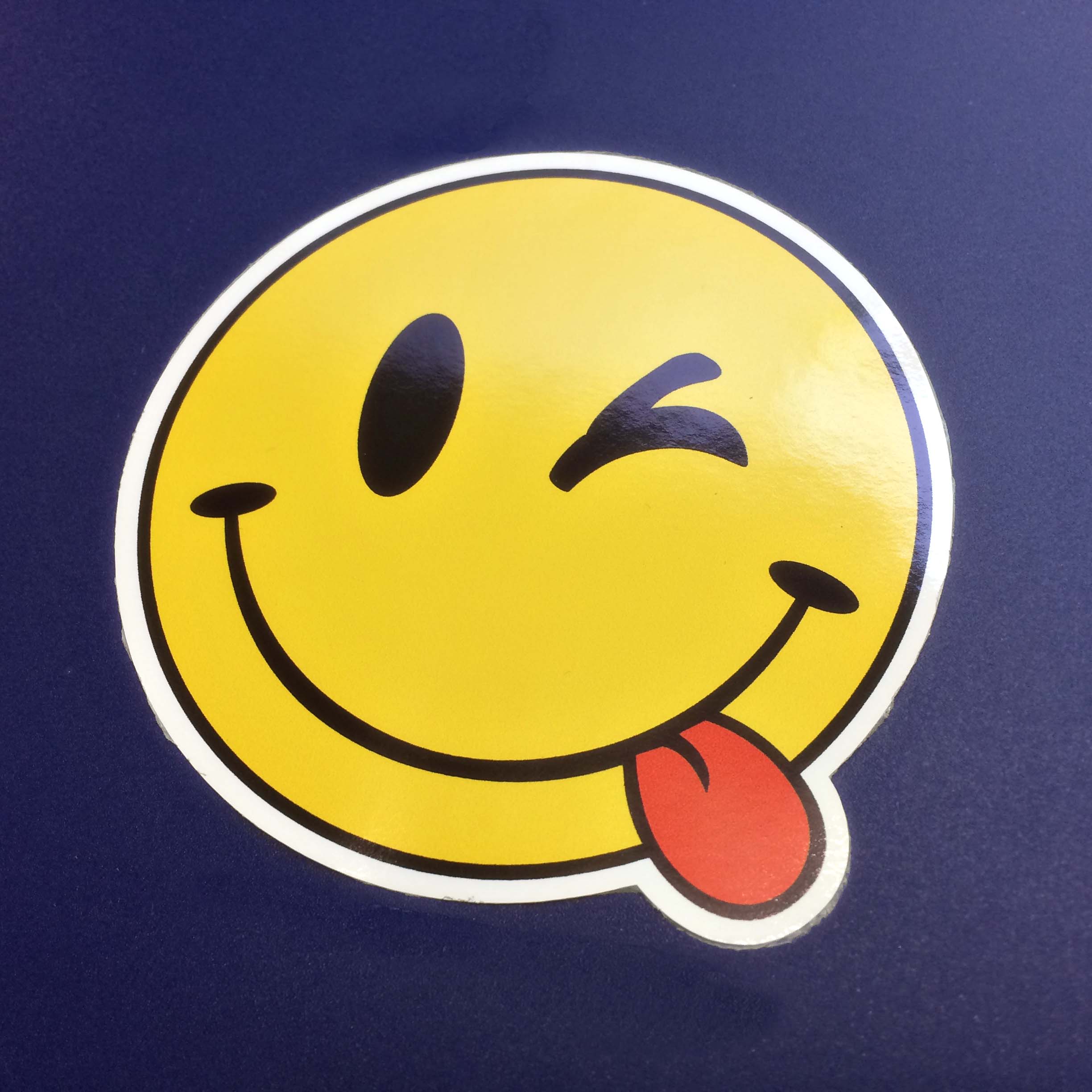 FLUORESCENT CHEEKY SMILEY EMOJI STICKER. A fluorescent round smiling face. One eye is winking and a red tongue is hanging out of the corner of the mouth.