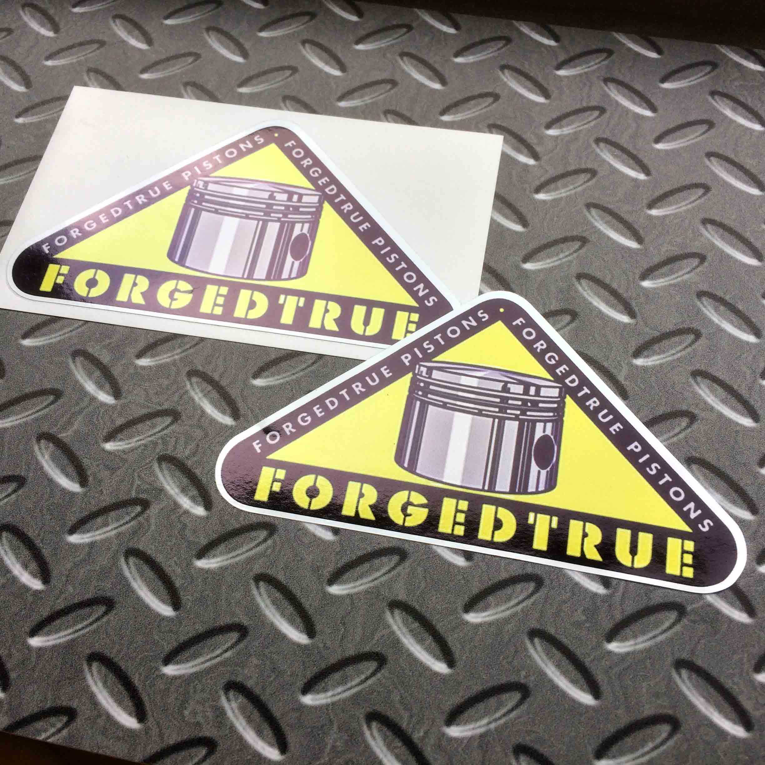FORGEDTRUE PISTONS VINTAGE STICKERS. A piston on a yellow triangle bordered in black. Forgedtrue Pistons in grey lettering is along two sides. Forgedtrue in bold yellow lettering is across the base of the triangle.