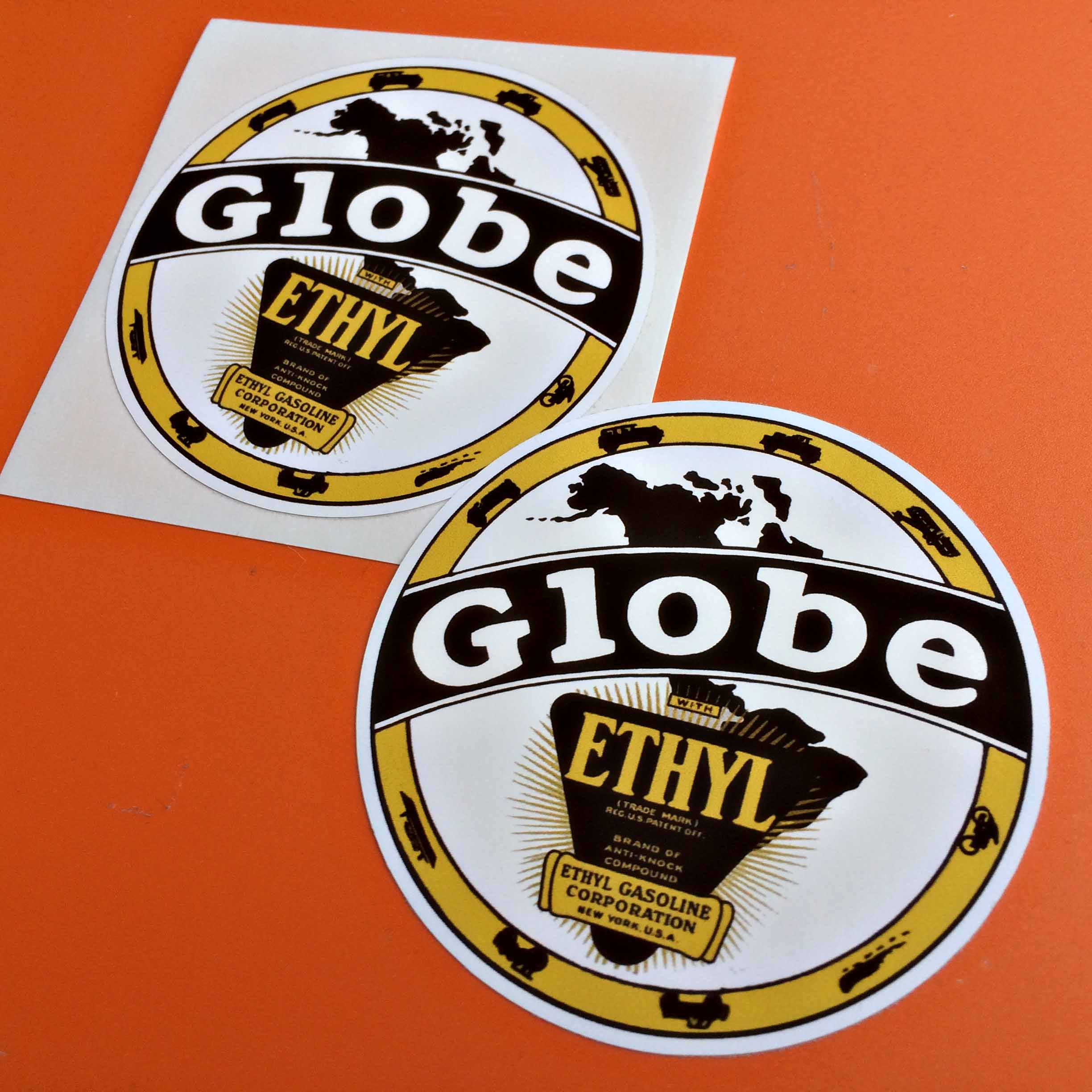GLOBE ETHYL GASOLINE STICKERS. Globe in white lettering on a black banner sits across tow concentric circles in yellow and white. In the centre is a map of the world and the Ethyl logo, a black inverted triangle with yellow lettering. Types of vehicles surround the outer circle.