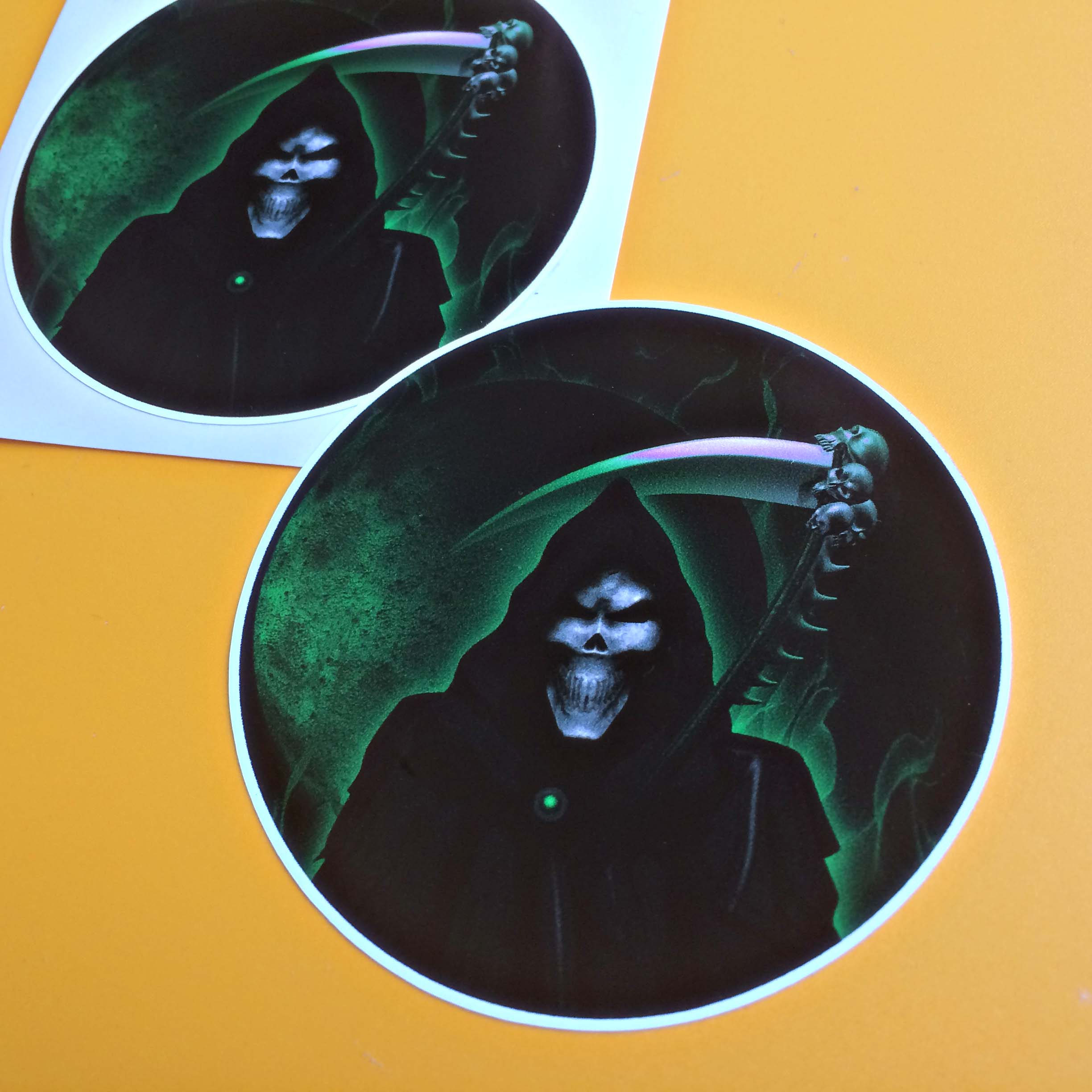 GRIM REAPER STICKERS. The grim reaper, a skeletal figure in a dark hooded robe carrying a scythe with skulls attached. The sky is a mist of black and green.