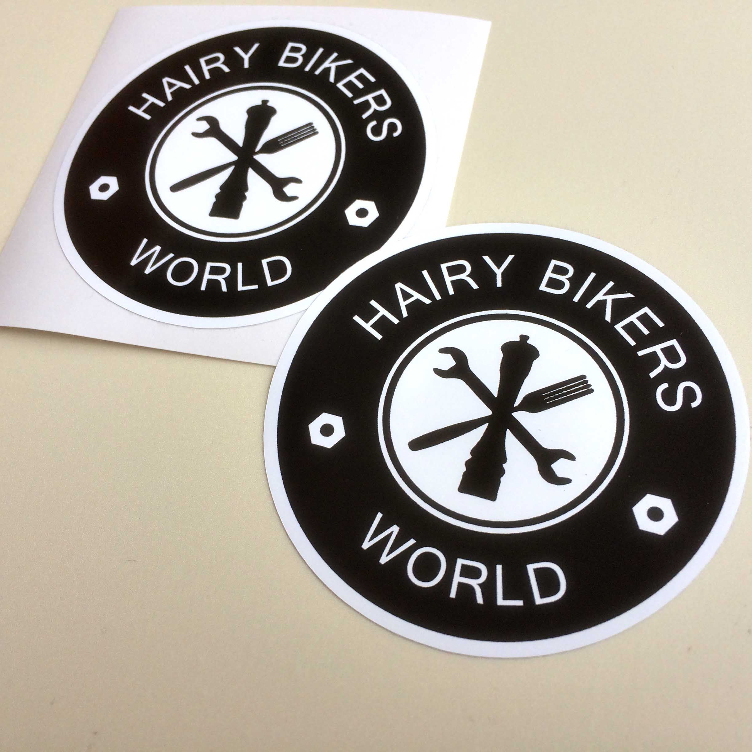 HAIRY BIKERS WORLD STICKERS. Two concentric circles in black and white. Hairy Bikers World in white lettering and two nuts on the outer circle. The white inner circle contains a spanner, a peppermill and a fork in black.