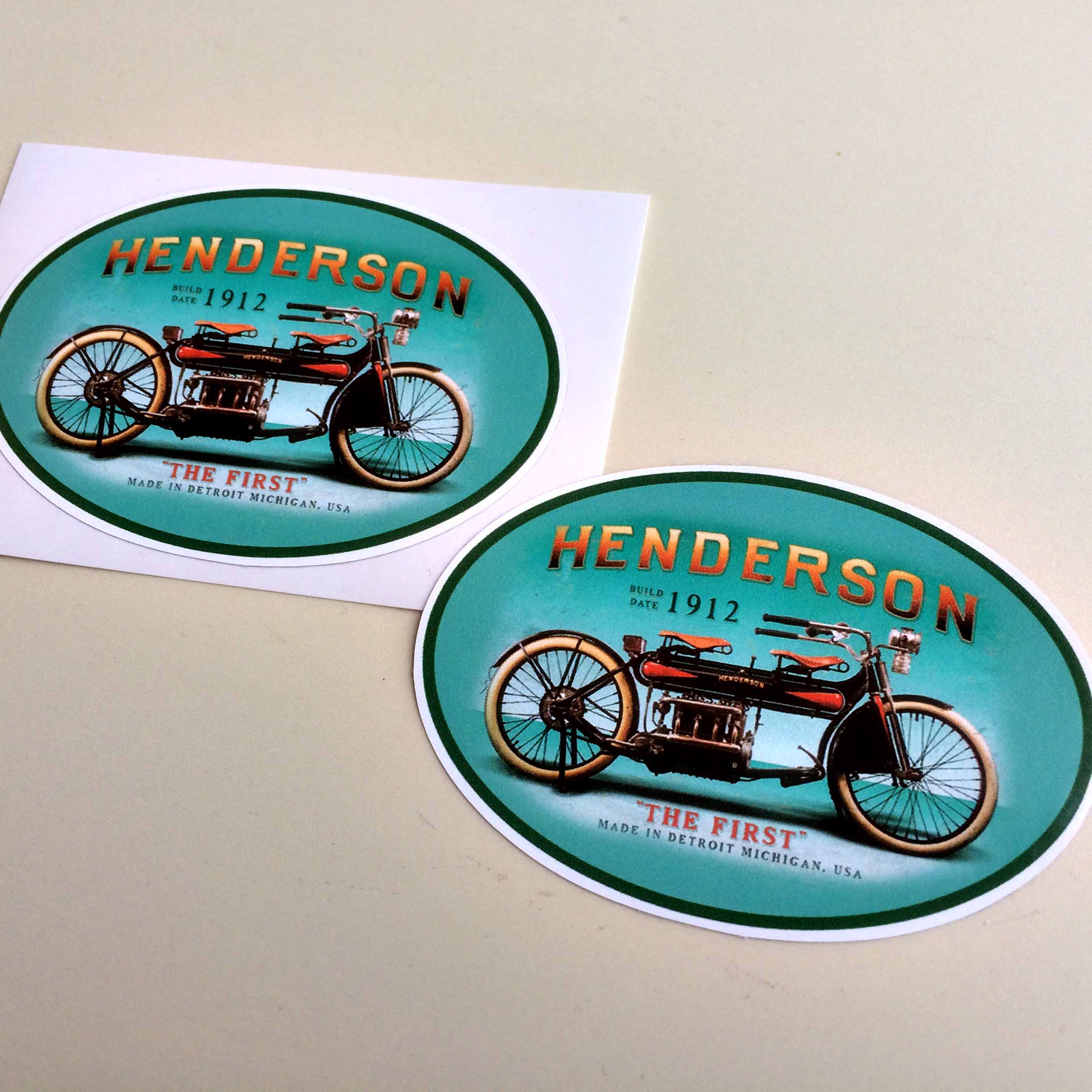 Henderson in gold and "The First" in red lettering surrounds a vintage motorcycle on a blue oval sticker. Additional text in black - Build Date 1912, Made in Detroit, Michigan, USA.