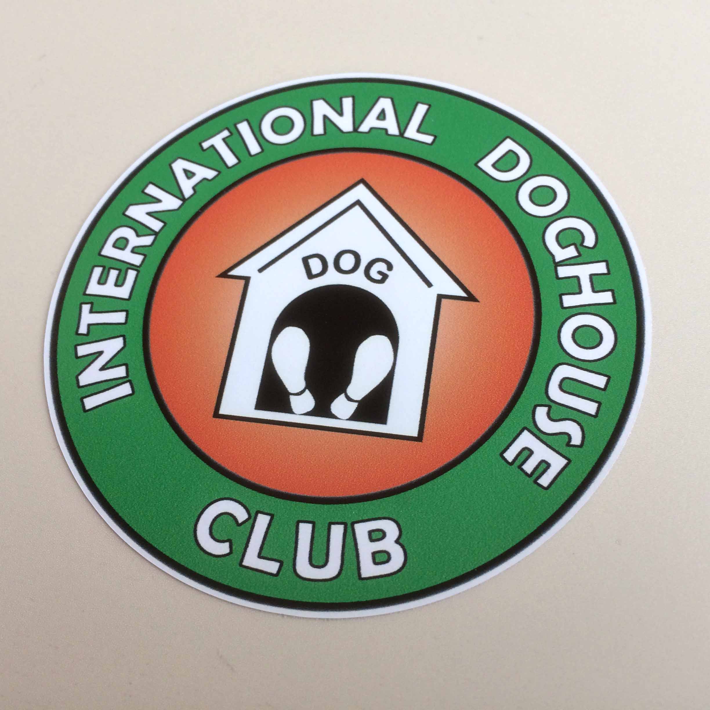 INTERNATIONAL DOGHOUSE CLUB STICKER. International Doghouse Club in white lettering on a green outer circle surrounds a white kennel on an orange background. The soles of two feet are in the doorway with the word Dog above it.