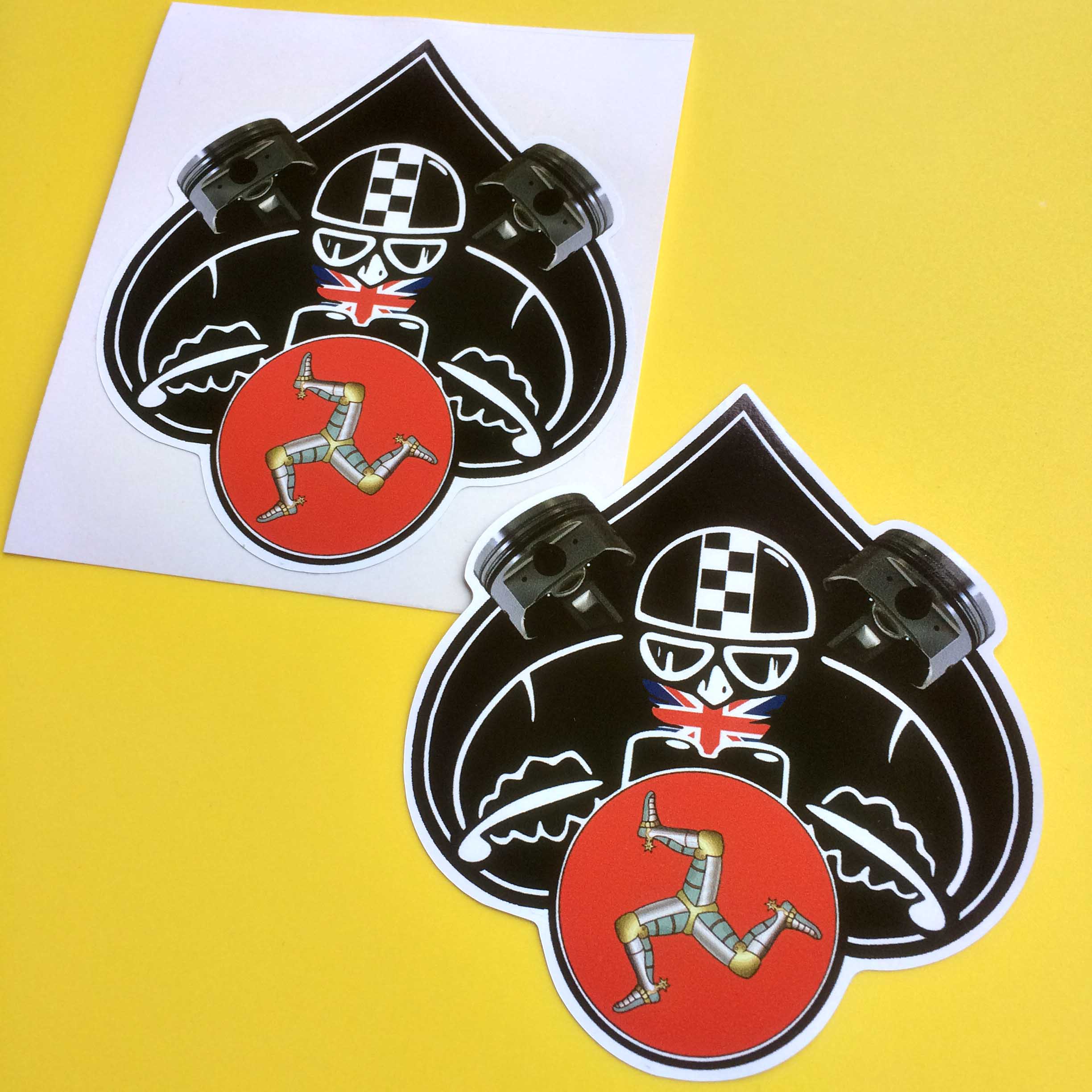 ISLE OF MAN CAFE RACER STICKERS. An inverted Ace of Spade above a circular Isle Of Man flag. Overlaying the Ace Of Spade are two piston heads and a motorcyclist wearing a chequered and Union Jack emblazoned helmet.