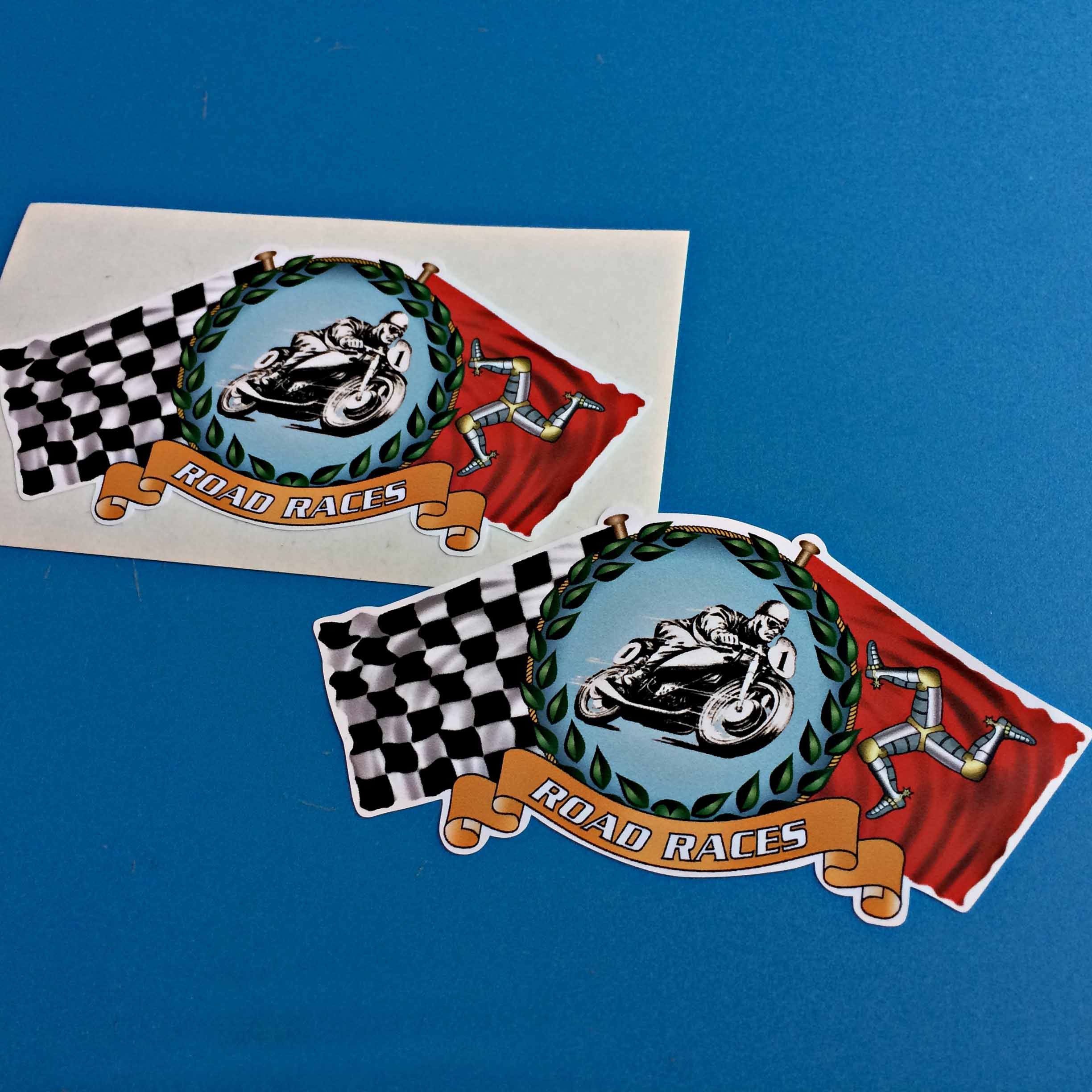 ISLE OF MAN CHEQUERED ROAD RACE STICKERS. Road Races in white lettering on a gold banner. Above is a man on a motorbike surrounded by a laurel wreath. Either side is a chequered flag and the Isle Of Man flag on poles.