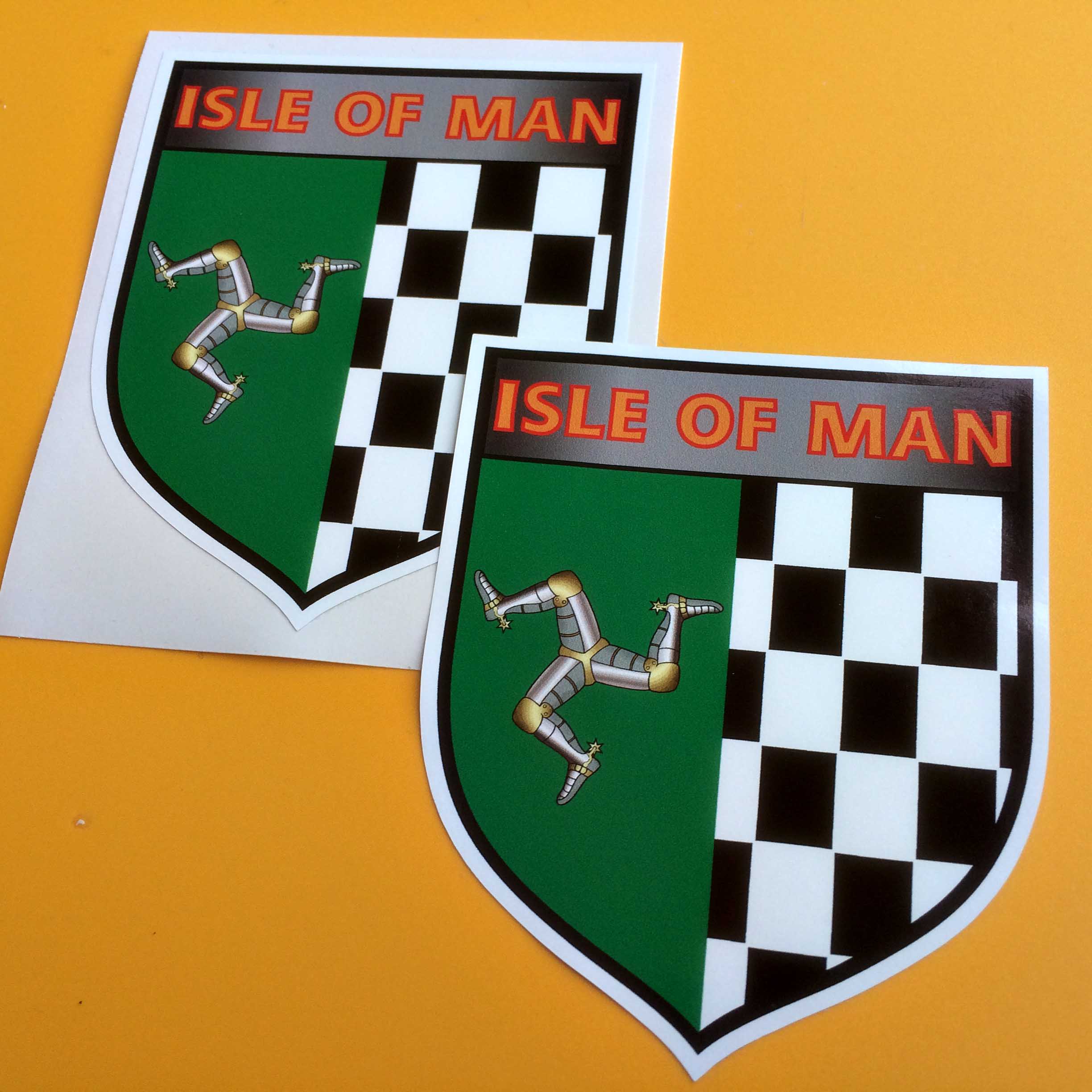 ISLE OF MAN CHEQUERED SHIELD STICKERS. A shield with a triskelion on a green background and black and white chequer. Isle Of Man in yellow and red lettering on a grey background is across the top of the shield.
