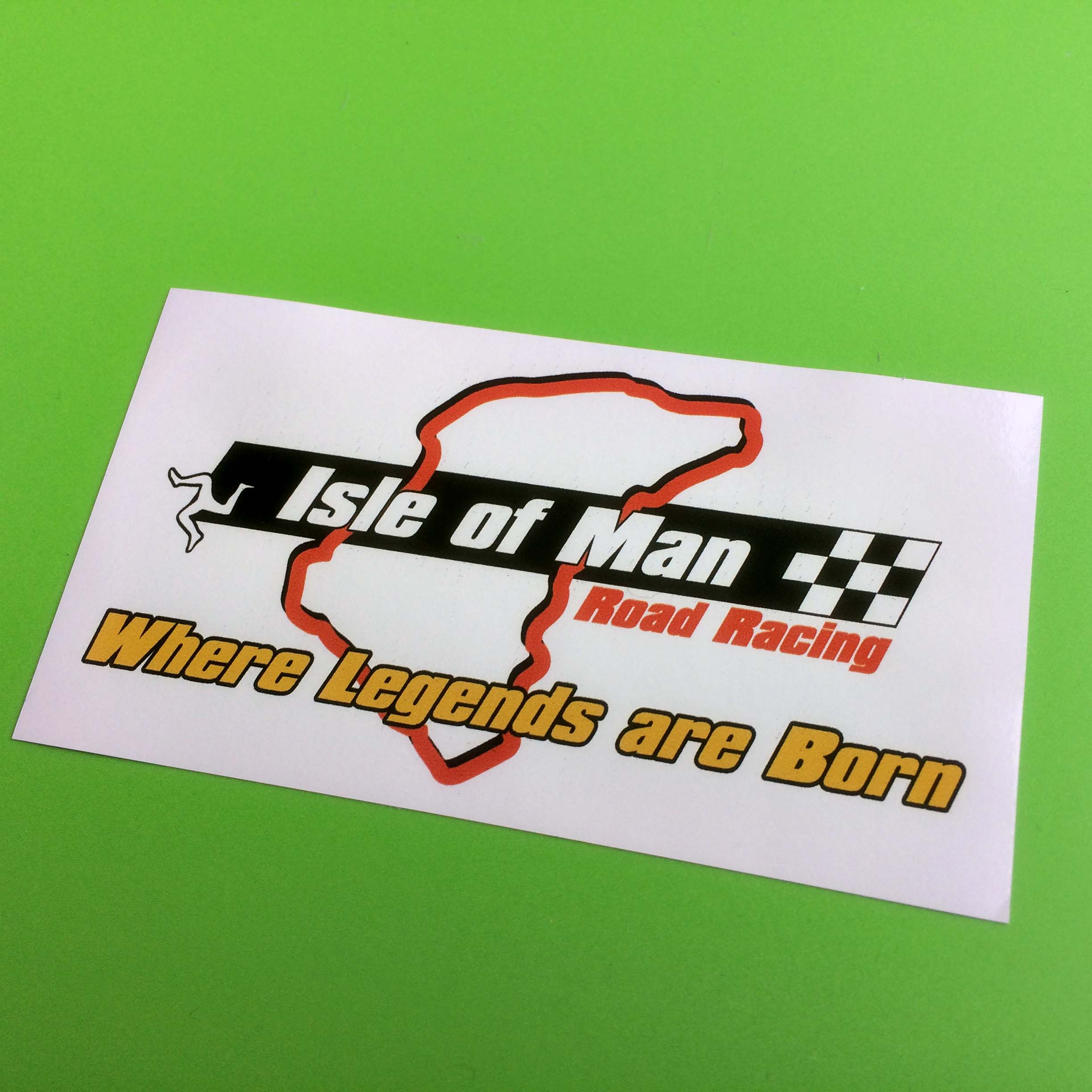 ISLE OF MAN ROAD RACING STICKER. Isle of Man in white lettering, a chequered flag and a white triskelion on a black banner. Below are Road Racing in red and Where Legends are Born in yellow lettering. A red contour of the race circuit is in the background.