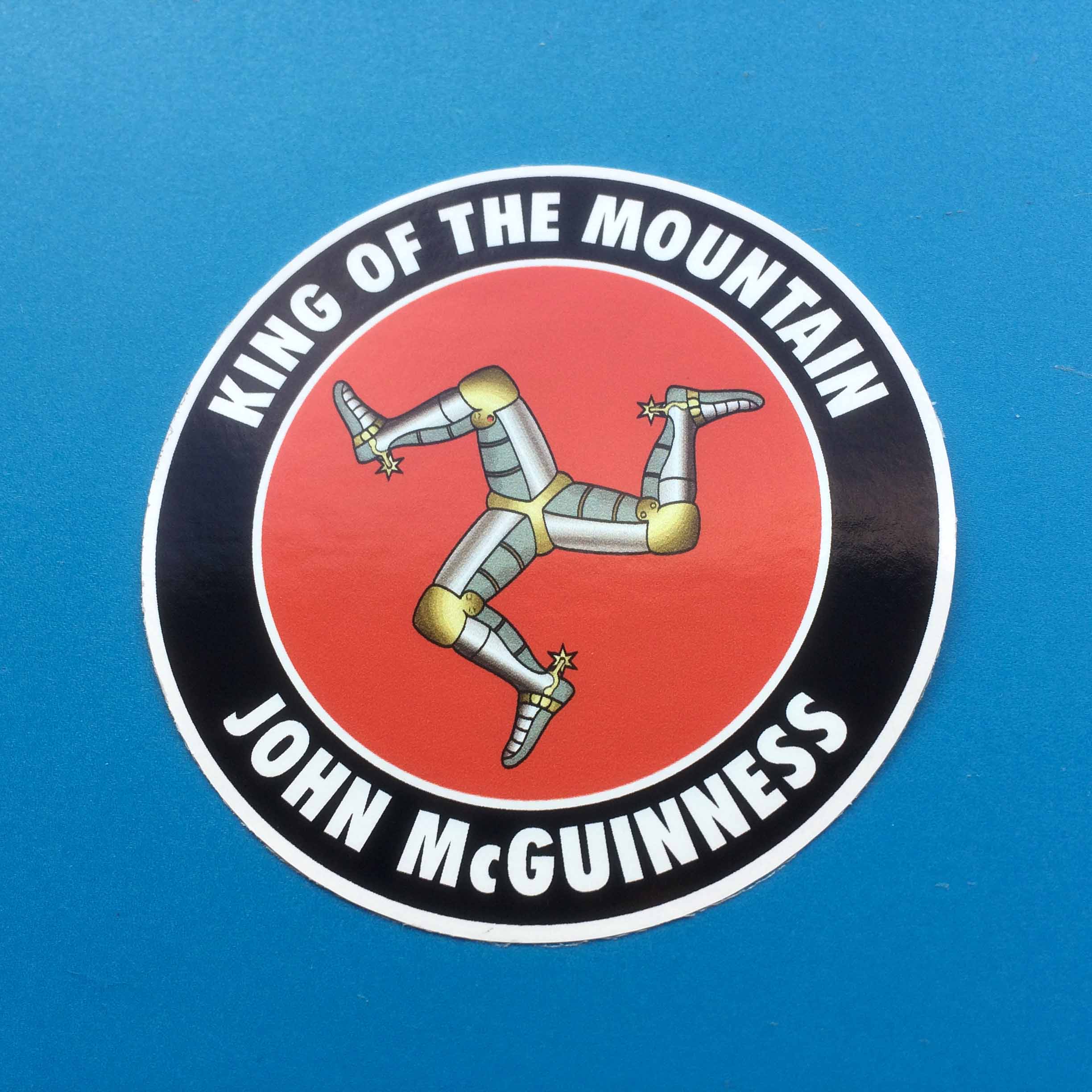 JOHN MCGUINESS KING OF THE MOUNTAIN STICKER. King Of The Mountain John McGuiness in white lettering on a black outer circle. A red inner circle contains three armoured legs with golden spurs.