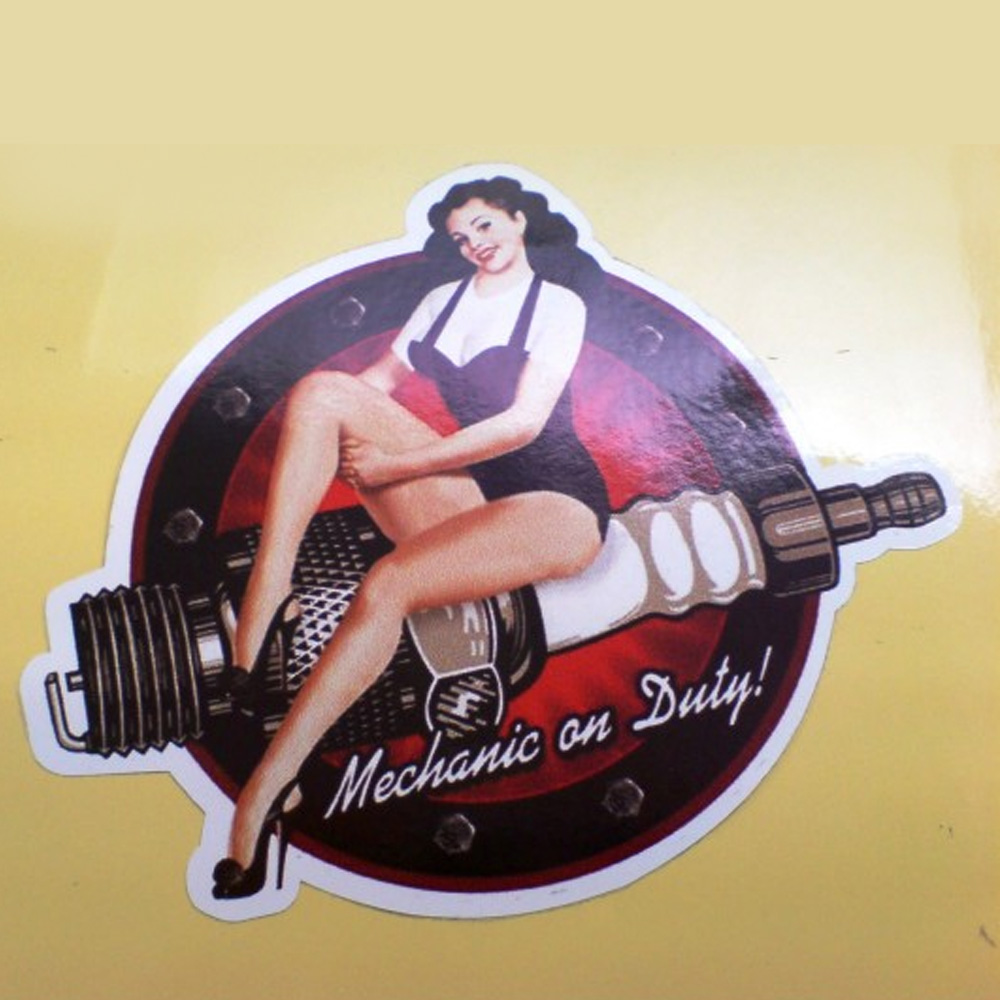 A black and red circular sticker. Mechanic on Duty! in white italic lettering and a model with long black hair wearing a black swimsuit and heels is sitting on a spark plug.