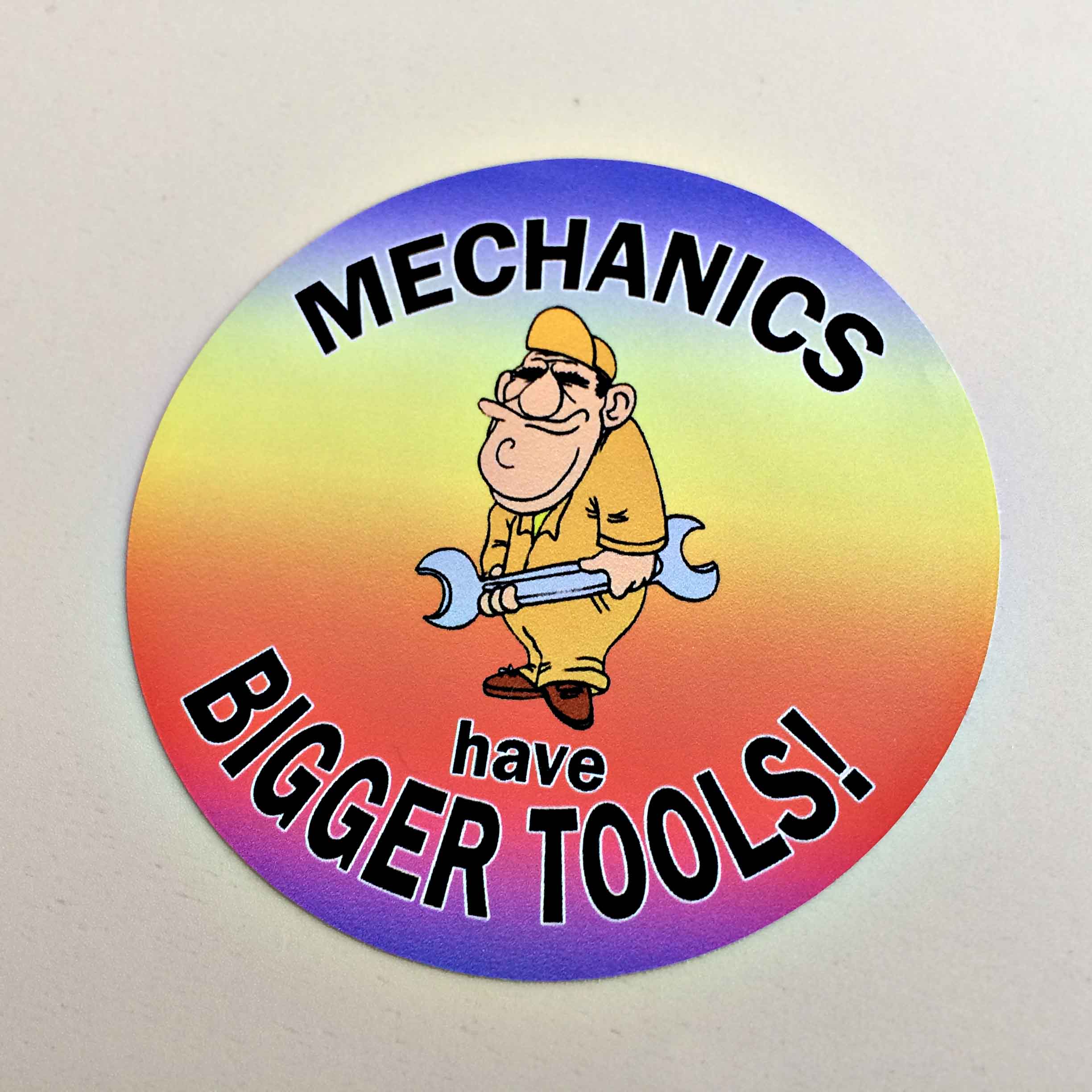MECHANICS HAVE BIGGER TOOLS STICKER COLOUR FADE. Humorous cartoon character of a man holding a spanner on a rainbow coloured background. With the wording Mechanics Have Bigger Tools surrounding the image.