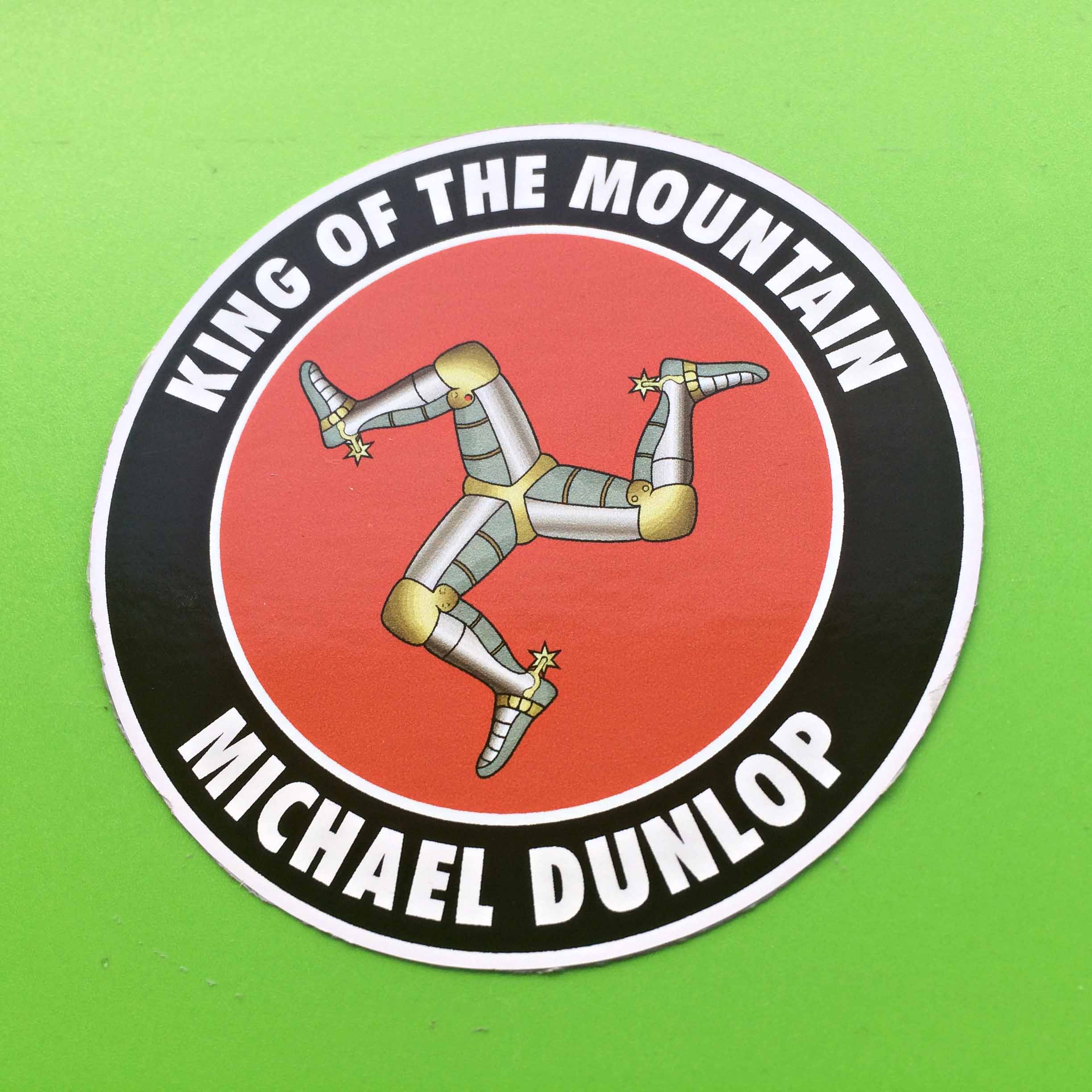 MICHAEL DUNLOP KING OF THE MOUNTAIN STICKER. King Of The Mountain Michael Dunlop in white lettering on a black outer circle. A red inner circle contains the three armoured legs with golden spurs of the Isle of Man.