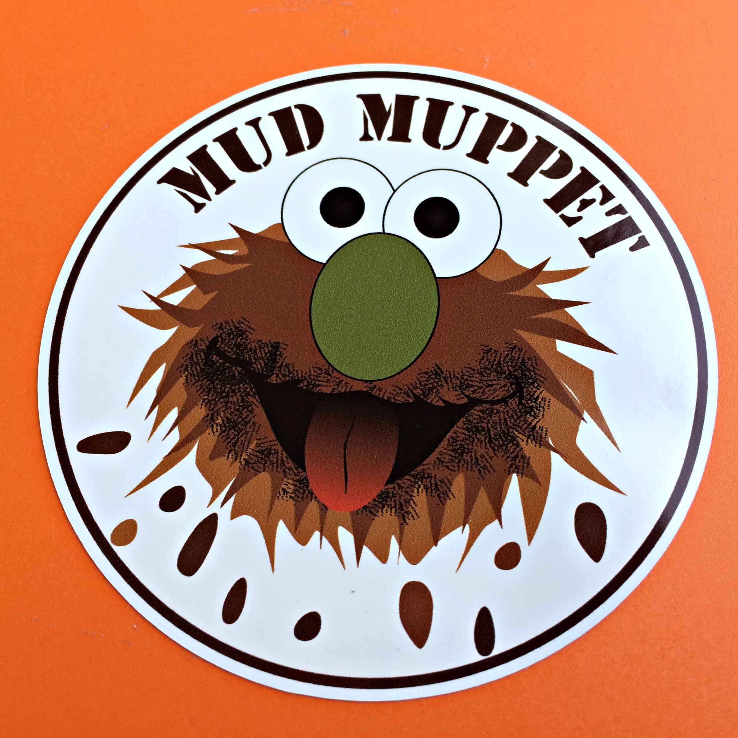 Mud Muppet in black lettering. A Muppet character with brown fur, a green nose, a red tongue and black and white eyes. Below are brown mud splatters.
