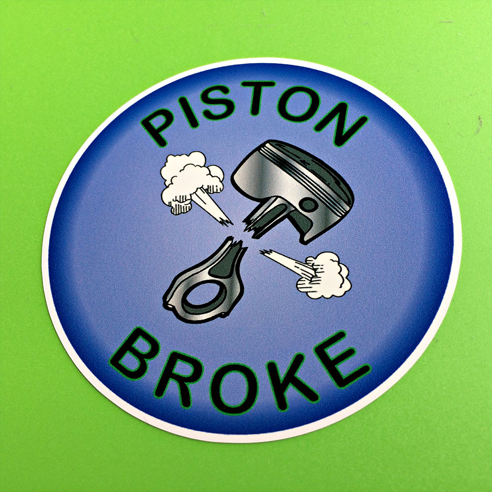 PISTON BROKE STICKER. A metal piston snapped in half with puffs of white smoke either side. Piston Broke surrounds the image in black lettering on a blue background.