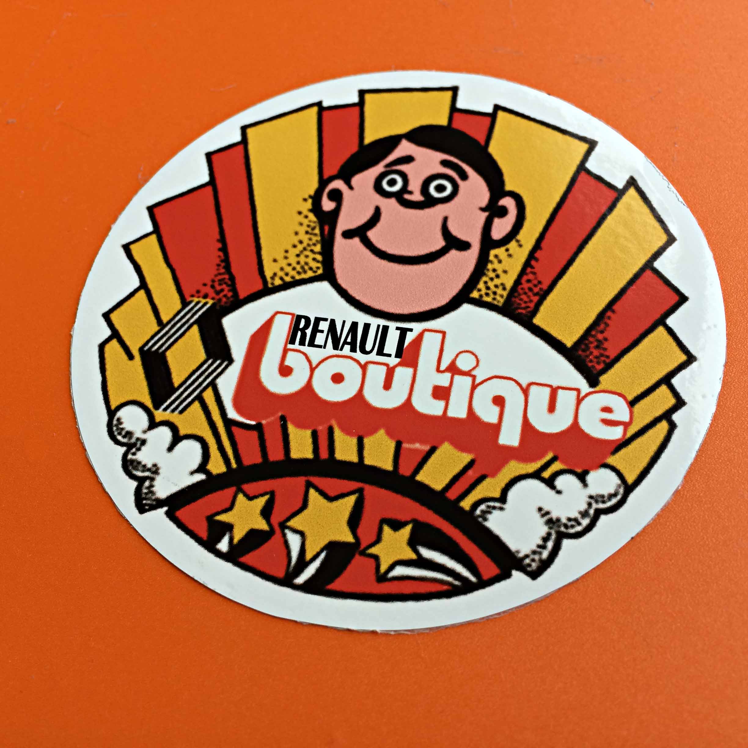RENAULT BOUTIQUE STICKER. A man with black hair smiling and wearing a white jumper with Renault boutique retro lettering and logo across it. Red and yellow columns of colour and three shooting stars surround the image.
