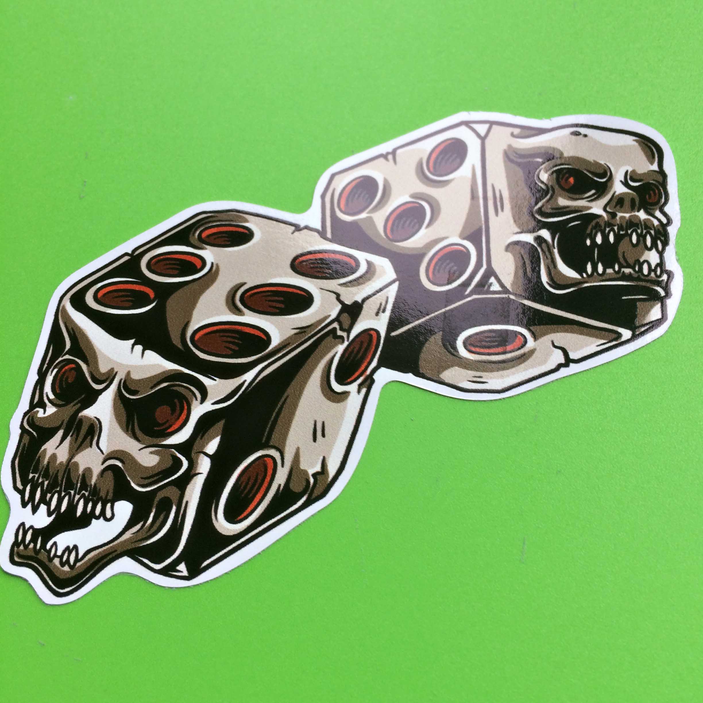 DEVILS DICE HOT ROD STICKER. Metallic looking twin dice with the dots in red. A skull with sharp teeth and red eye sockets is on the side of each dice.