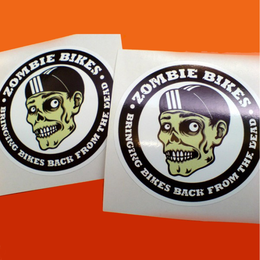 Zombie Bikes Bringing Bikes Back From The Dead in white lettering on black surrounds this circular sticker. In the centre on a white background is a green skull wearing a black cap with three white lines down the middle.