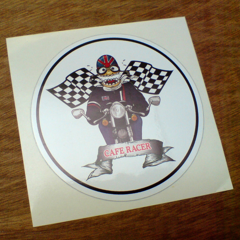 Cafe Racer in red lettering across a silver banner on a white circular sticker. Above is a cartoon character sat astride a vintage motorcycle. He is wearing goggles and a Union Jack helmet, blue jumper and jeans. His eyes and mouth are wide open. Chequered crossed flags are in the background.