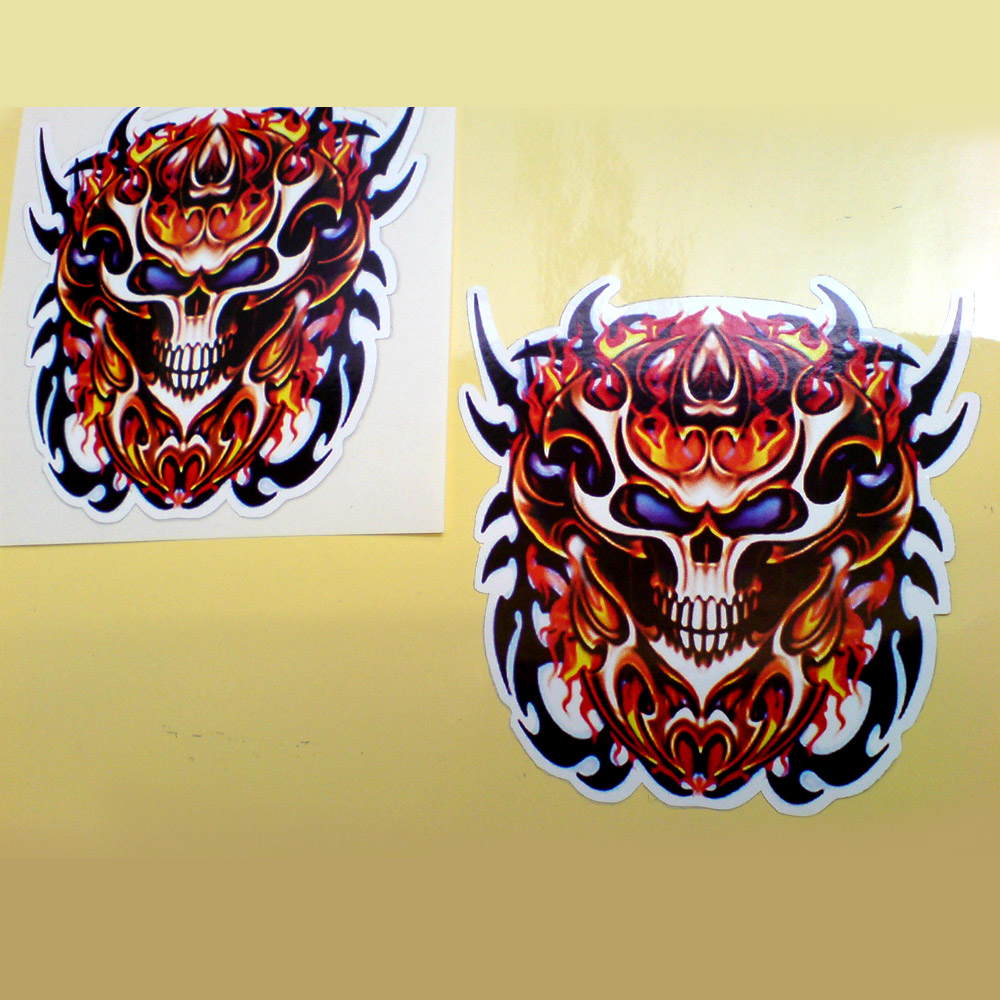 SKULL FIRESTORM STICKERS. A menacing looking skull with blue eyes surrounded by red, yellow and orange flames and black, sharp, jagged edges.