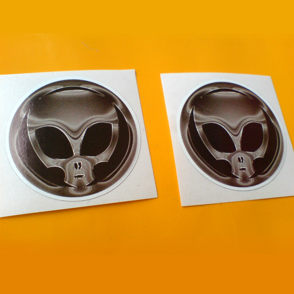 A metallic skull with black eyes on a black background. A circular sticker with a metallic border.