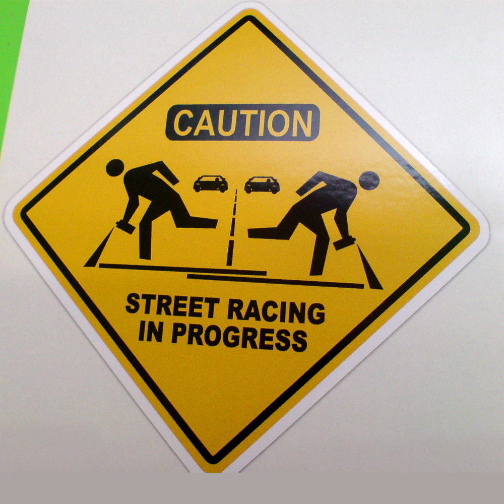 STREET RACING IN PROGRESS STICKER. Caution Street Racing In Progress on a yellow and black diamond sticker. Two cars and two men with spray cans are both sides of the road.