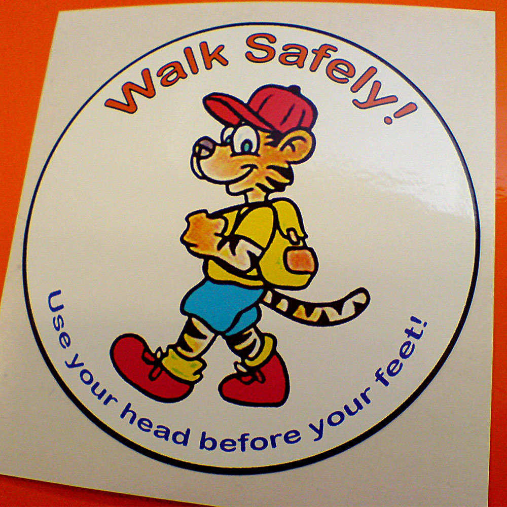 WALK SAFELY STICKER. Walk Safely! in red Use your head before your feet! in blue lettering surrounds a humorous tiger wearing blue shorts, red boots and cap, a yellow shirt and carrying a backpack on a white circular sticker.