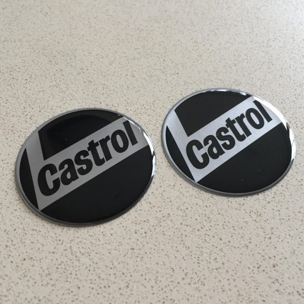 CASTROL STICKERS DOMED RESIN GEL. Castrol in black lowercase lettering on a silver L shaped background. Chrome edged. Black background.