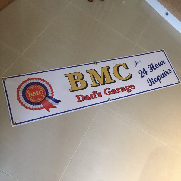 BMC 24 HOUR REPAIRS BANNER. On the left is the red, white and blue BMC rosette. Centre is BMC bold gold lettering. Dad's Garage lettering in red below. Right in blue lettering is For 24 Hour Repairs. White banner with blue edging.