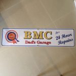 On the left is the red, white and blue BMC rosette. Centre is BMC bold gold lettering. Dad's Garage lettering in red below. Right in blue lettering is For 24 Hour Repairs. White banner with blue edging.